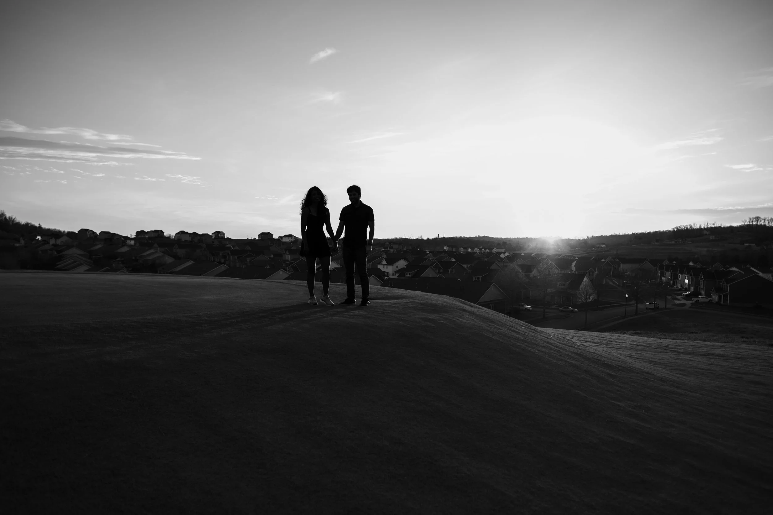 Silhouette of a couple holding hands on a grassy hill at sunset, with a neighborhood and sky in the background.