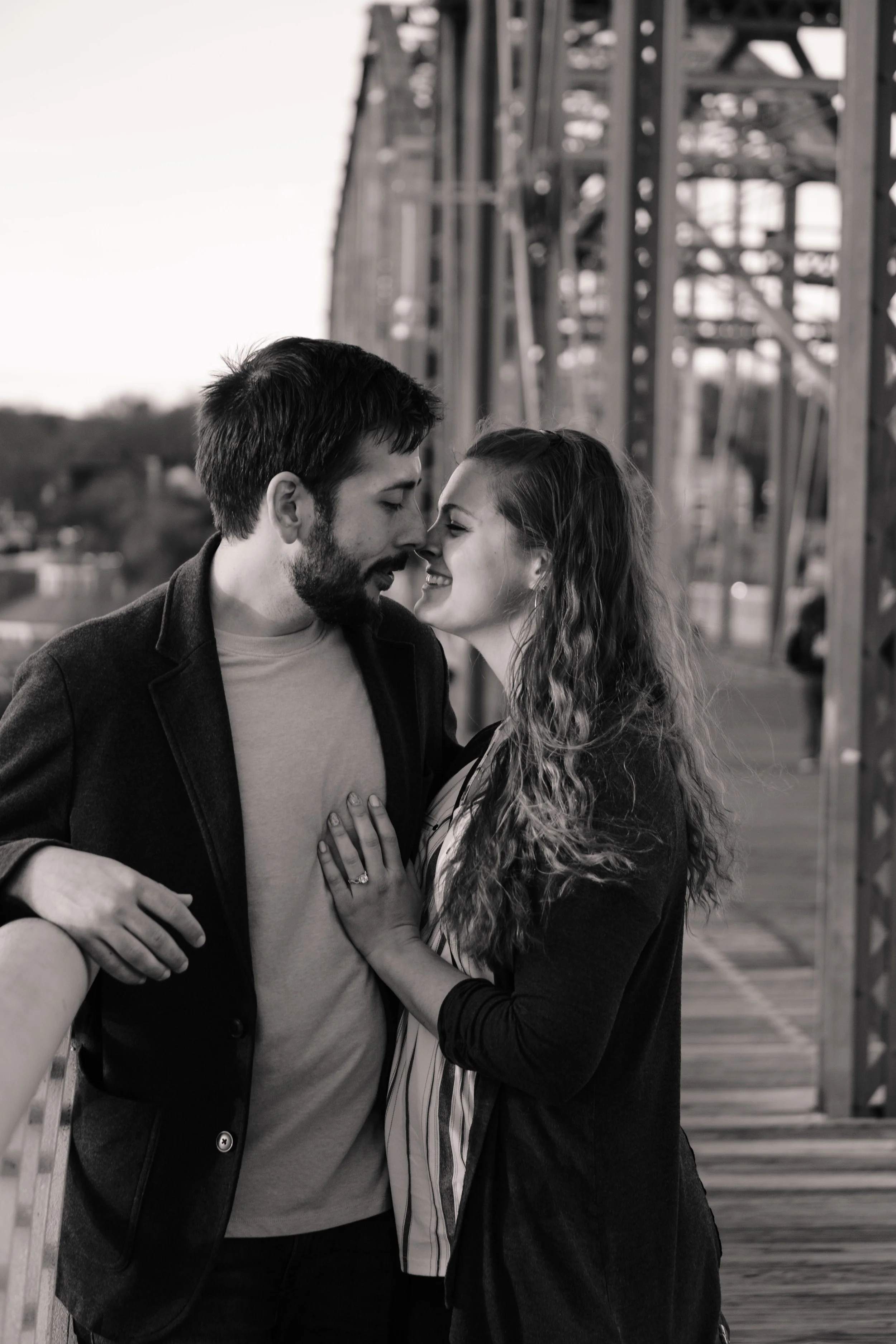 Black-and-white photo of a couple with foreheads touching and smiling on a bridge, with their hands on each other's chests, surrounded by a metal bridge structure.