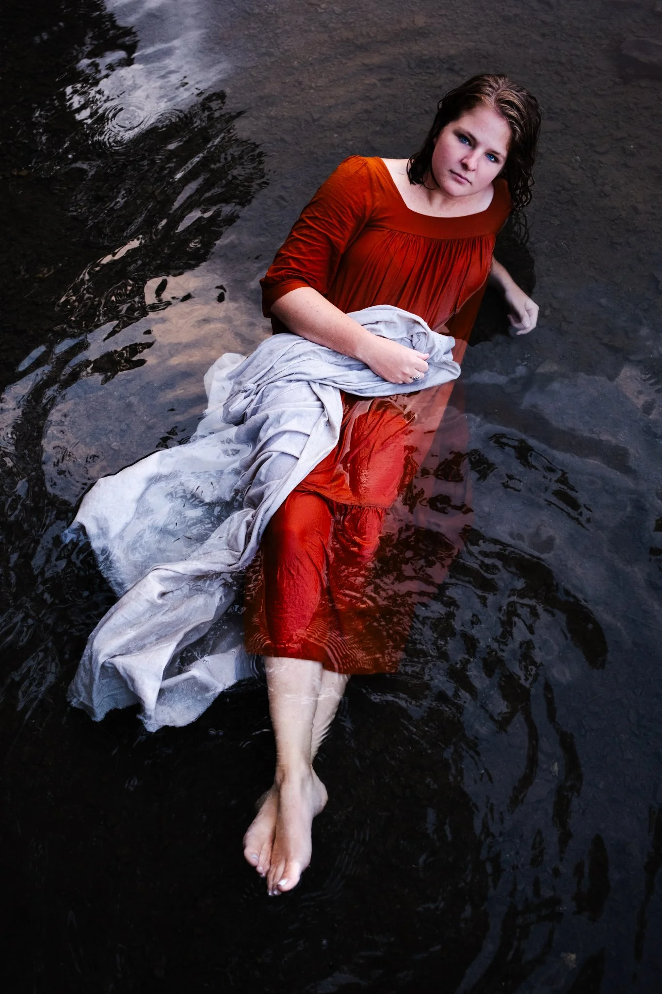 A woman with brown hair, wearing a red dress, sitting in dark water with a white cloth over her lap, gazing up at the camera.