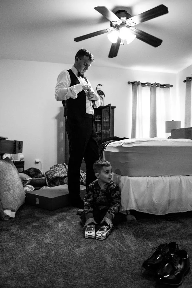 A man in formal attire adjusting his shirt or vest while a young boy sits on the floor with toy cars in front of him, inside a bedroom with a bed, window, and ceiling fan.