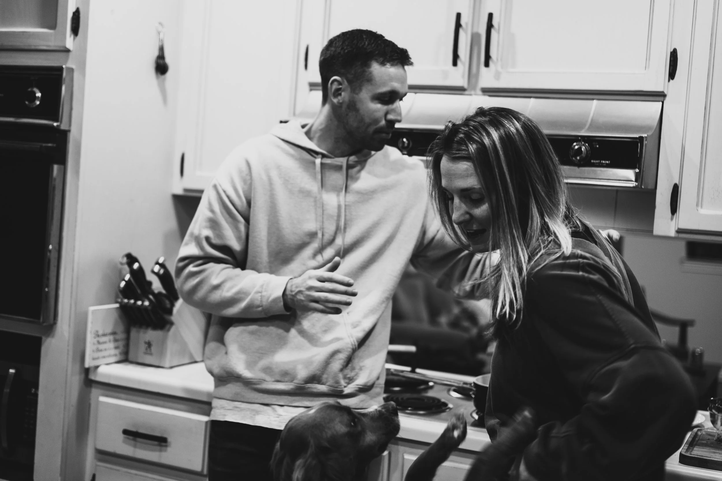 A man and woman in a kitchen, engaged with a dog. The man is wearing a light hoodie, and the woman has long hair and is wearing a dark jacket. The dog is being held by the woman, reaching up towards them.