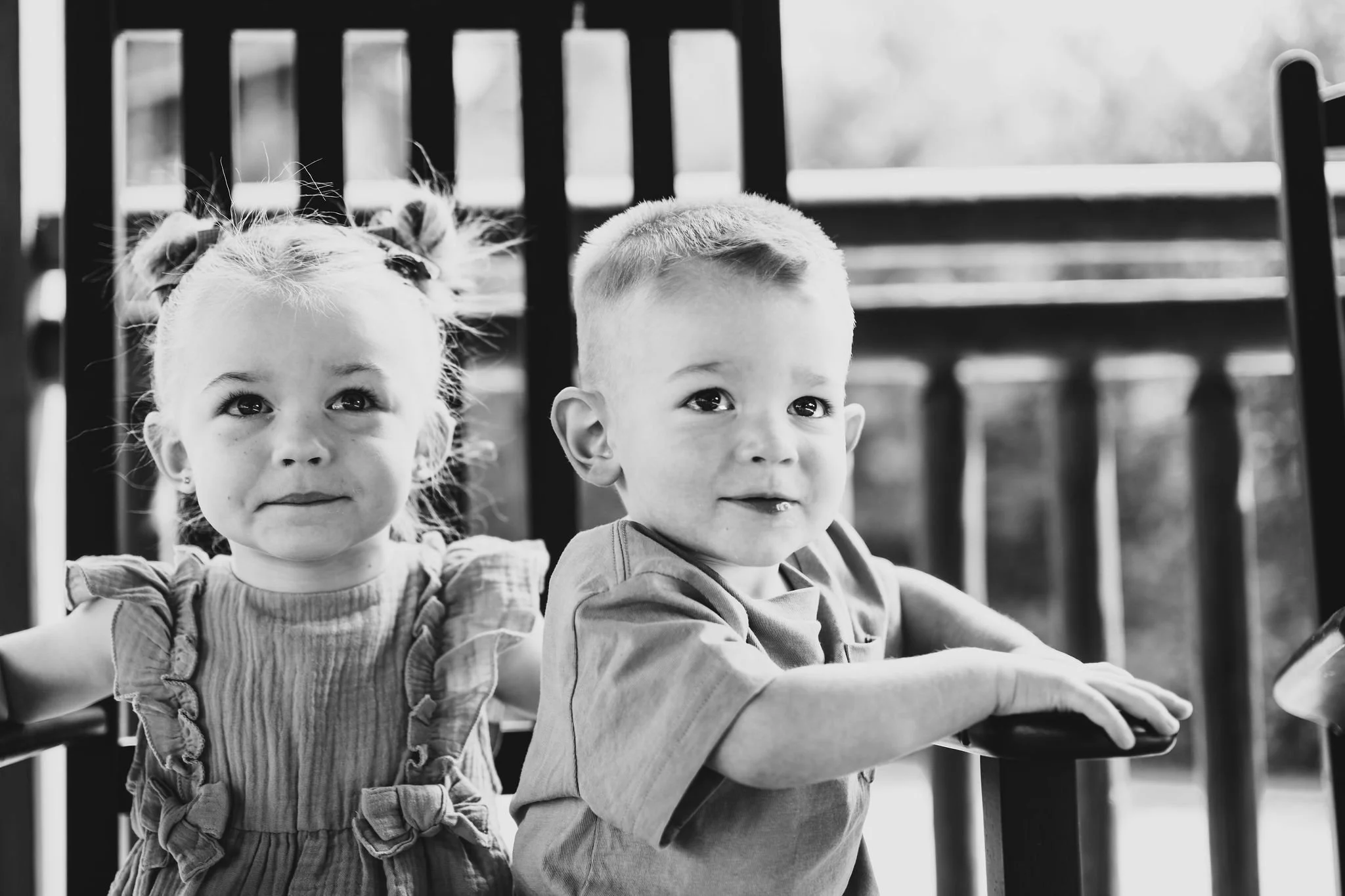 Black and white photo of a young girl and boy, sitting next to each other, looking at the camera. The girl has her hair in pigtails, wearing a dress with ruffles, and appears to be smiling slightly. The boy has short hair, wearing a T-shirt, and is s