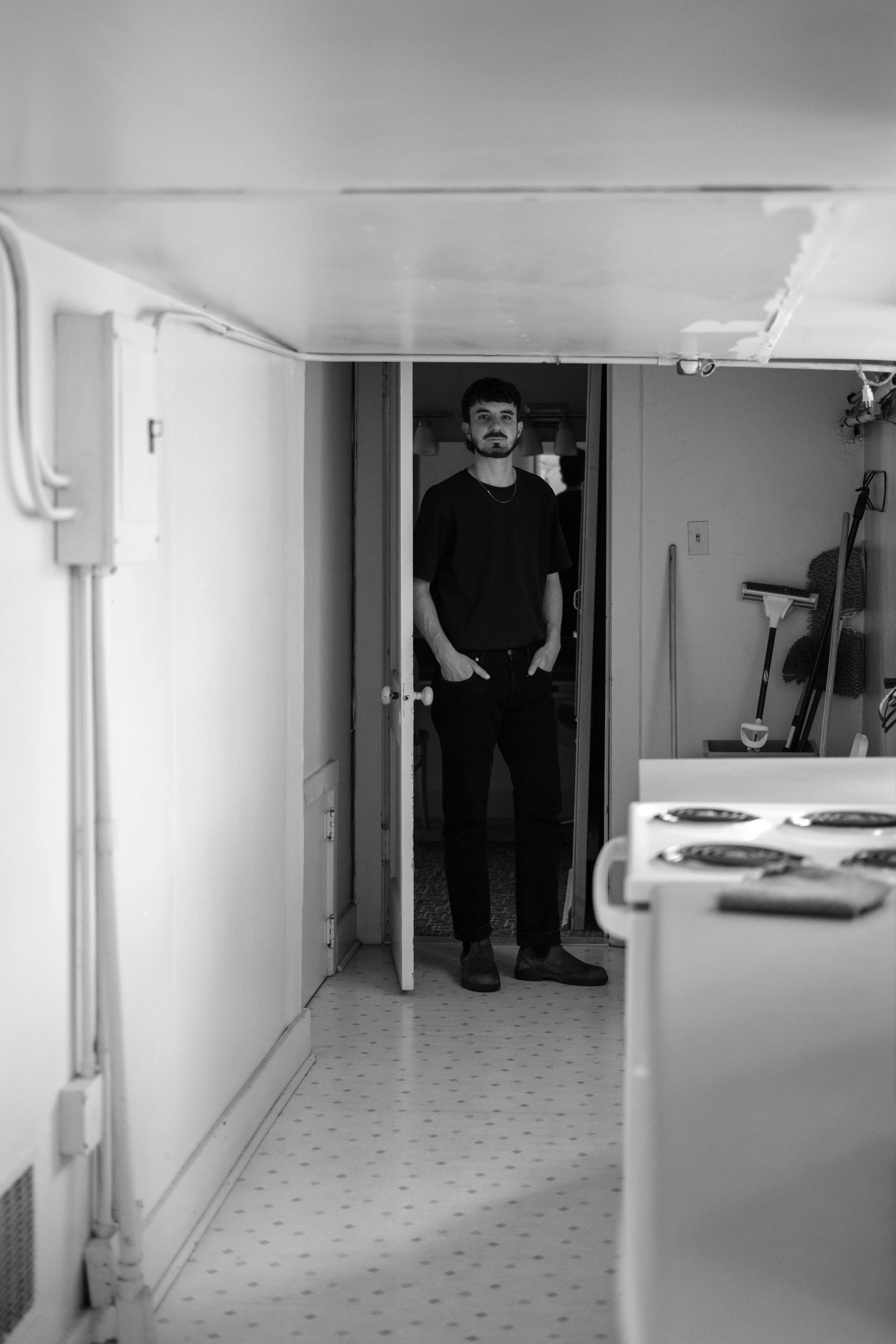 A man with a beard and dark hair, dressed in a black t-shirt and black pants, standing in a kitchen doorway.