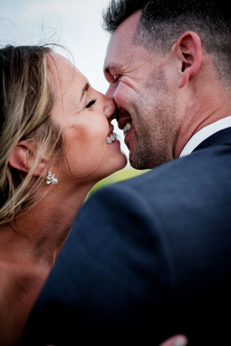 A close-up of a couple smiling and touching noses, with the woman wearing pearl earrings and the man in a suit, outdoors.