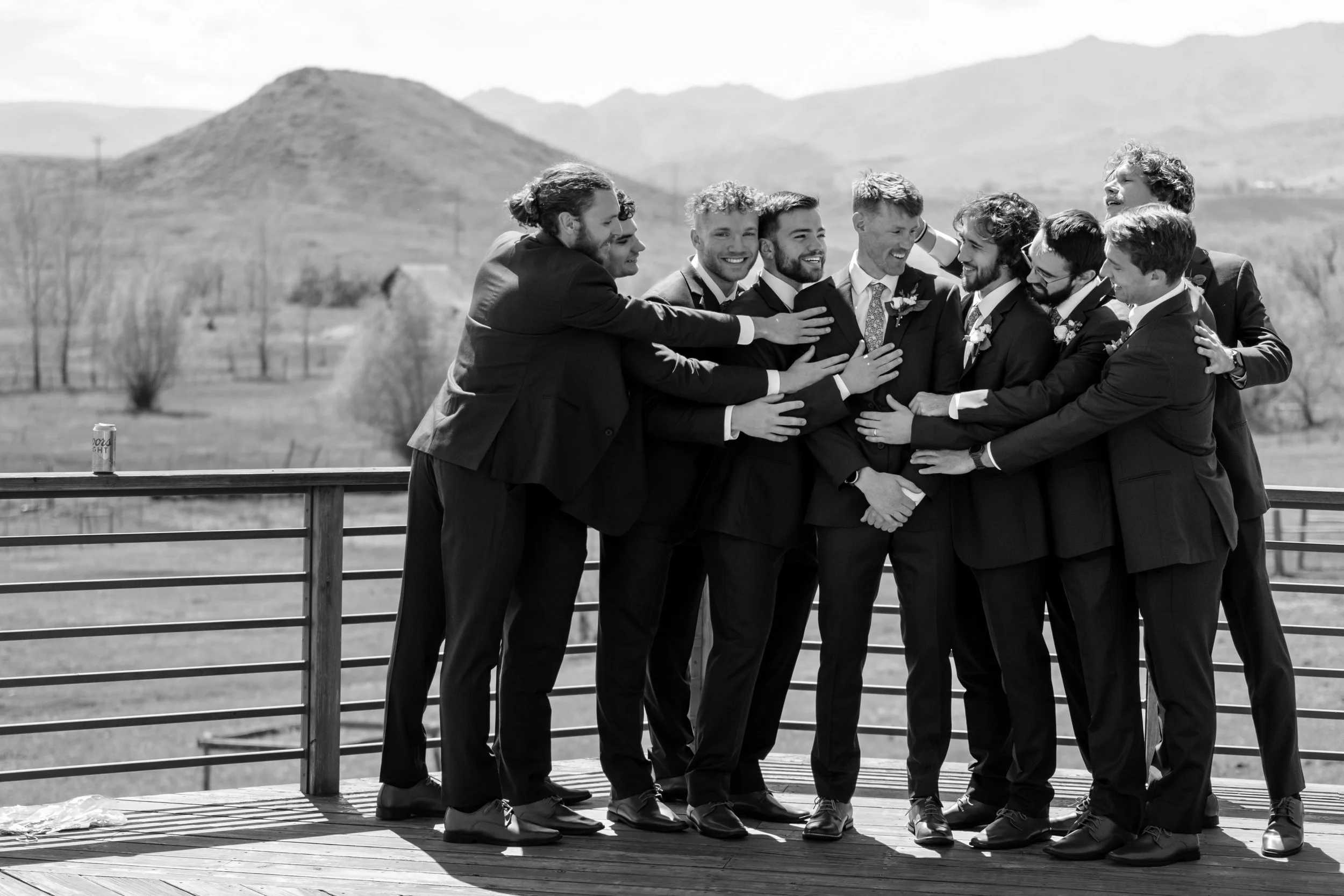 A group of men in tuxedos hugging and laughing outdoors on a balcony with mountains in the background, celebrating a wedding.