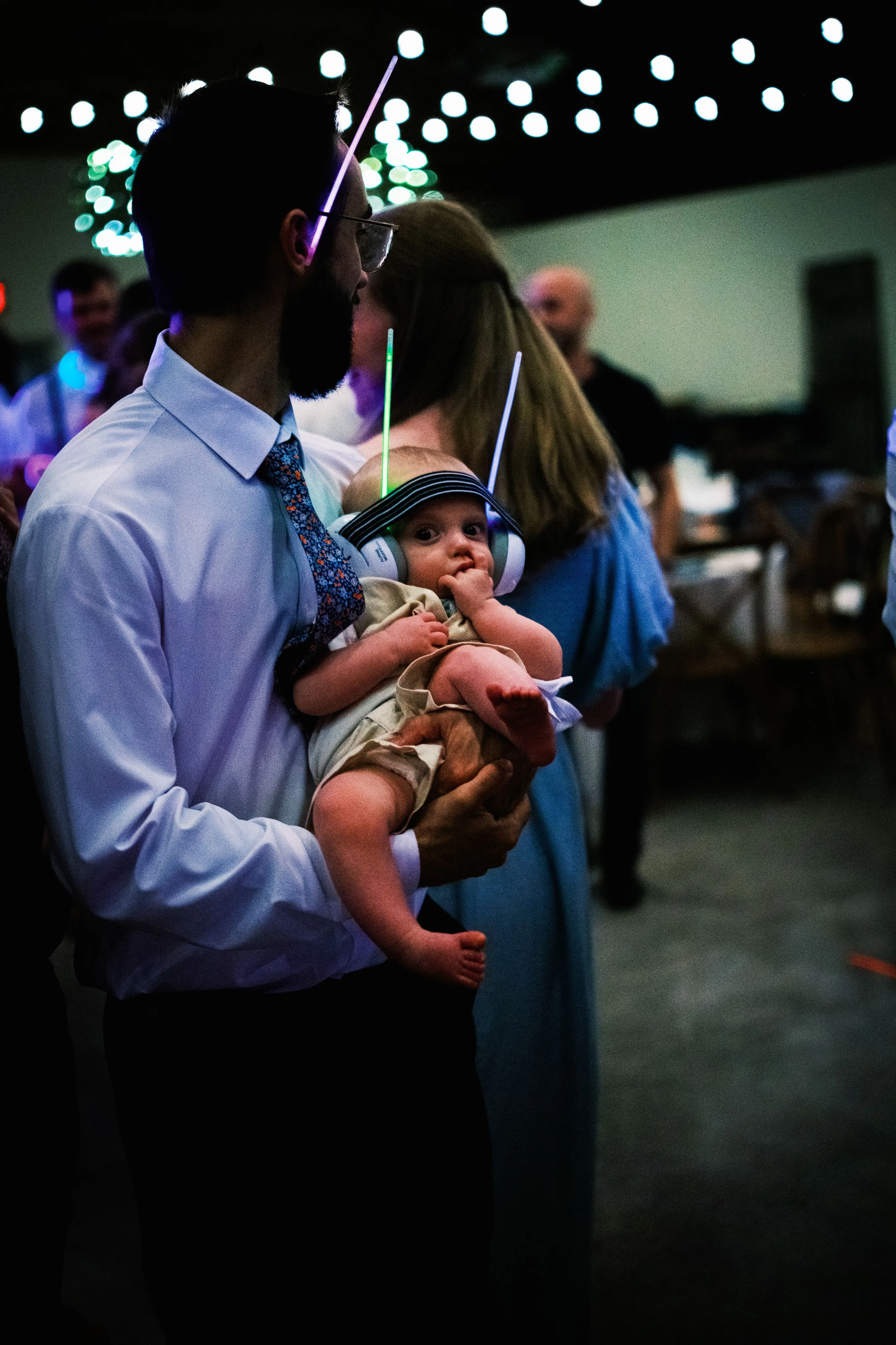 Man holding a baby at a party or event with string lights hanging from the ceiling in the background.