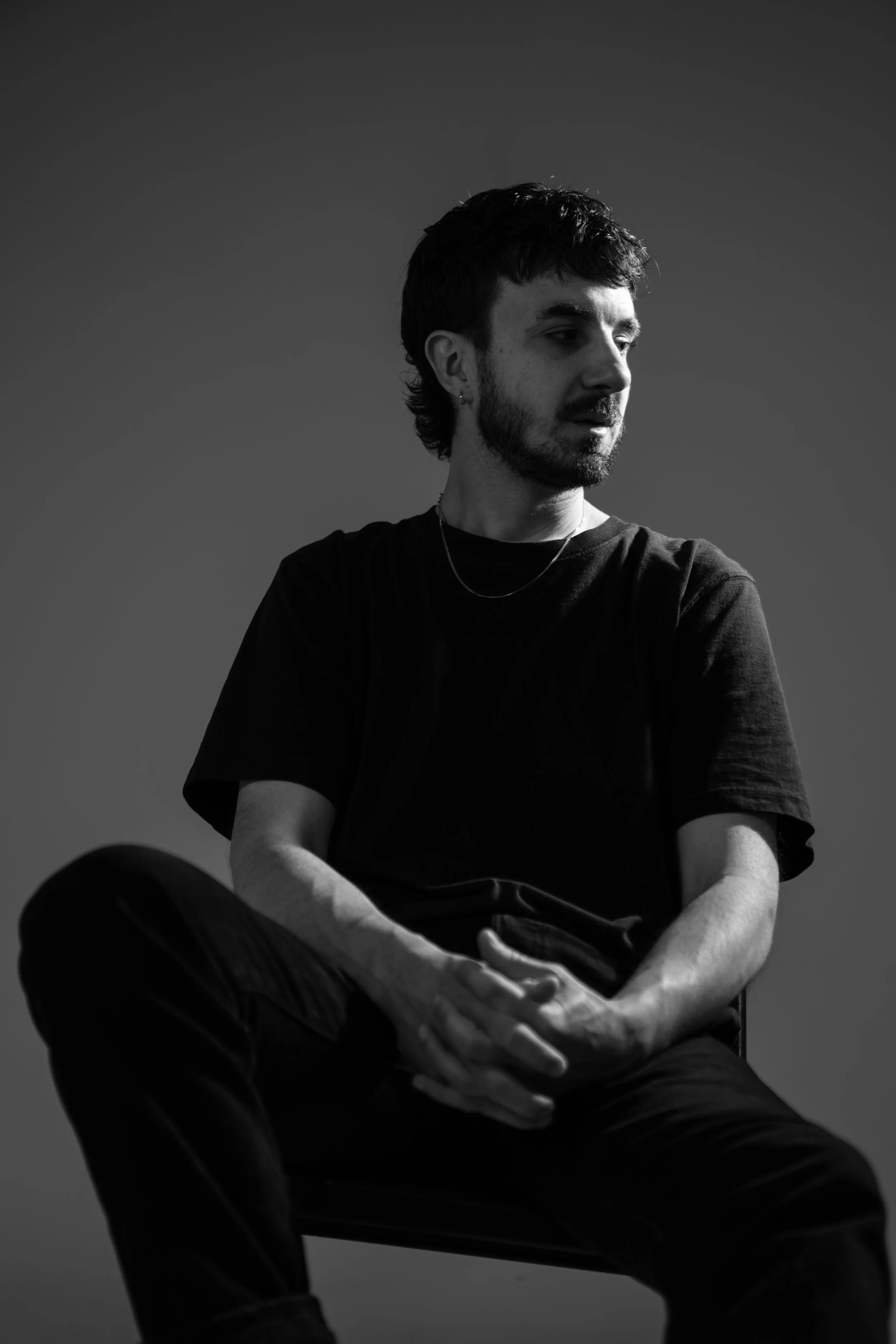 A young man with dark hair and beard, wearing a black t-shirt and a silver necklace, sitting on a chair against a plain background.
