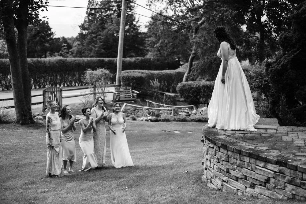 A woman in a long white dress standing on a brick platform, looking down at a group of women in dress standing on the grass, watching her.
