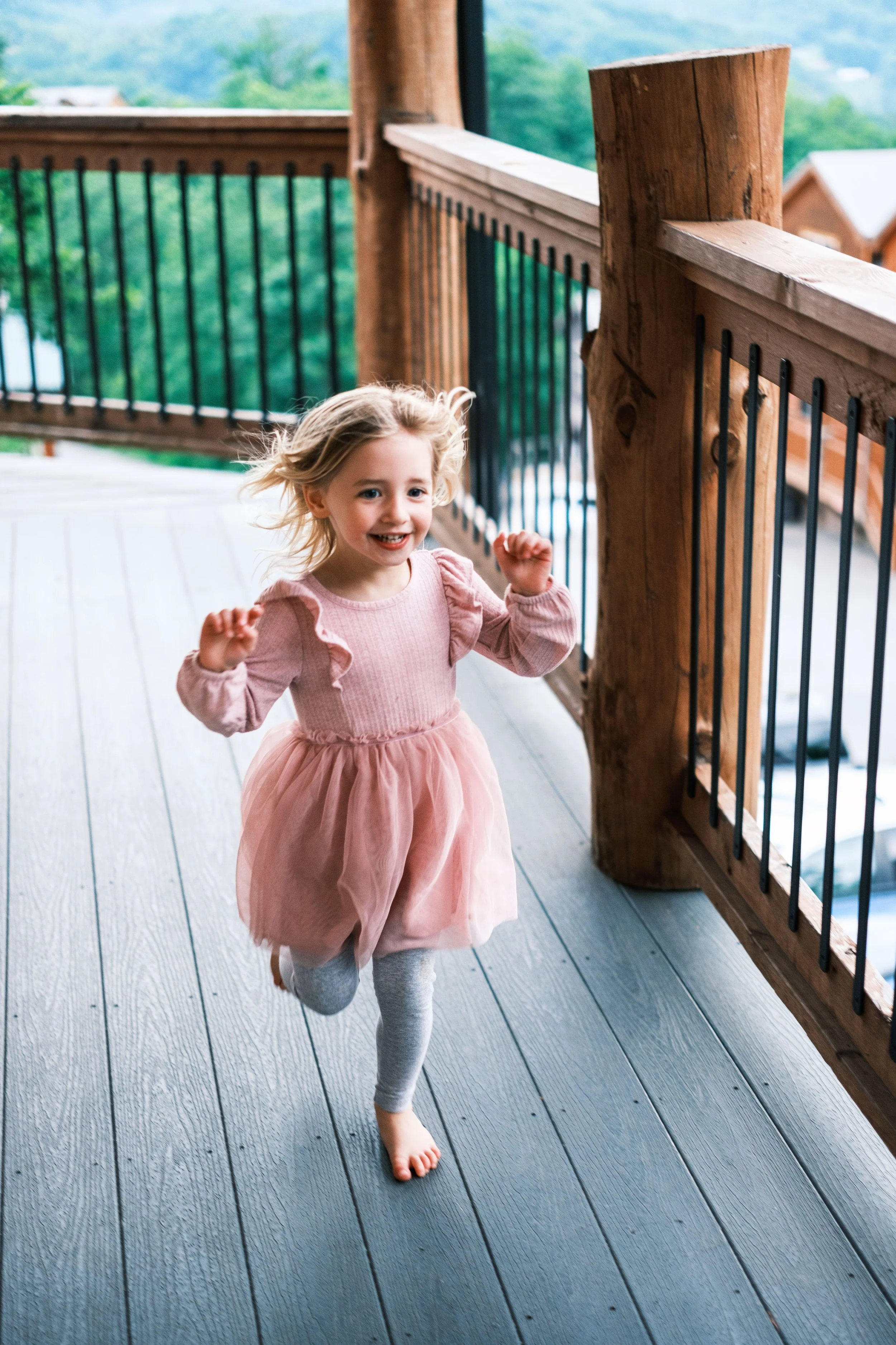 A young girl running on a porch with a wooden railing, smiling and wearing a pink dress with puffy sleeves and gray leggings.