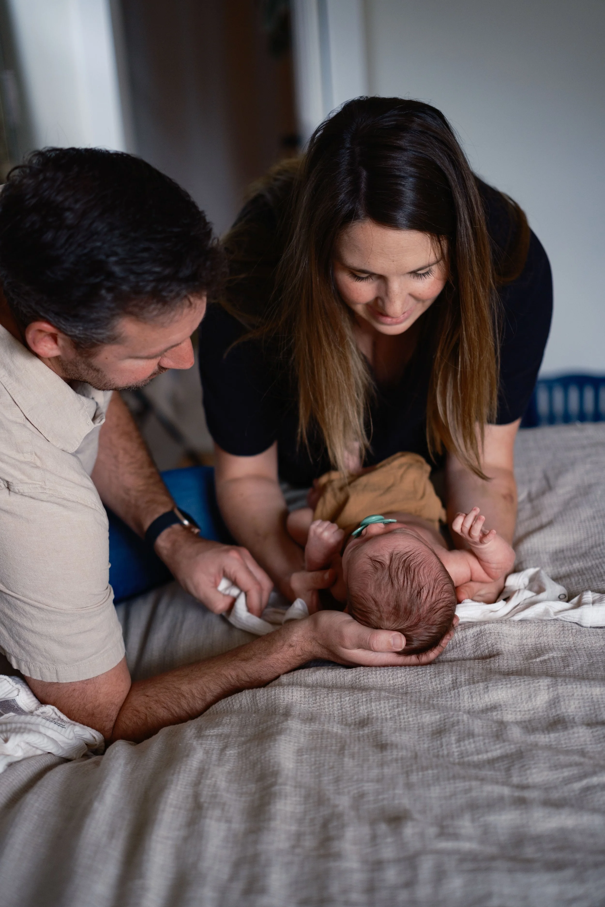 A family with a father, mother, and newborn baby in a home setting, with the mother and father gently holding the baby on a bed.