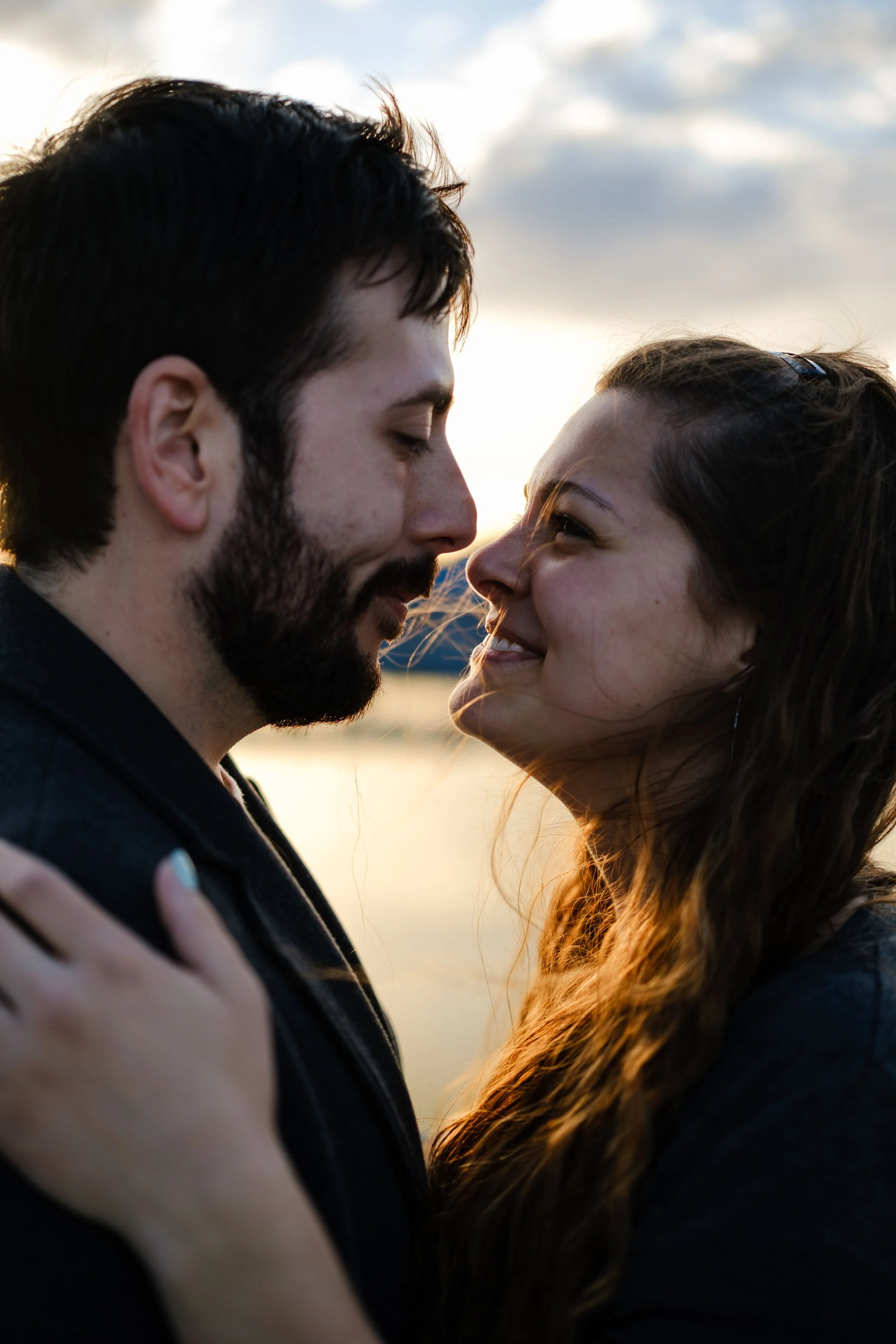 A young couple standing close together outdoors at sunset, smiling and looking into each other's eyes.