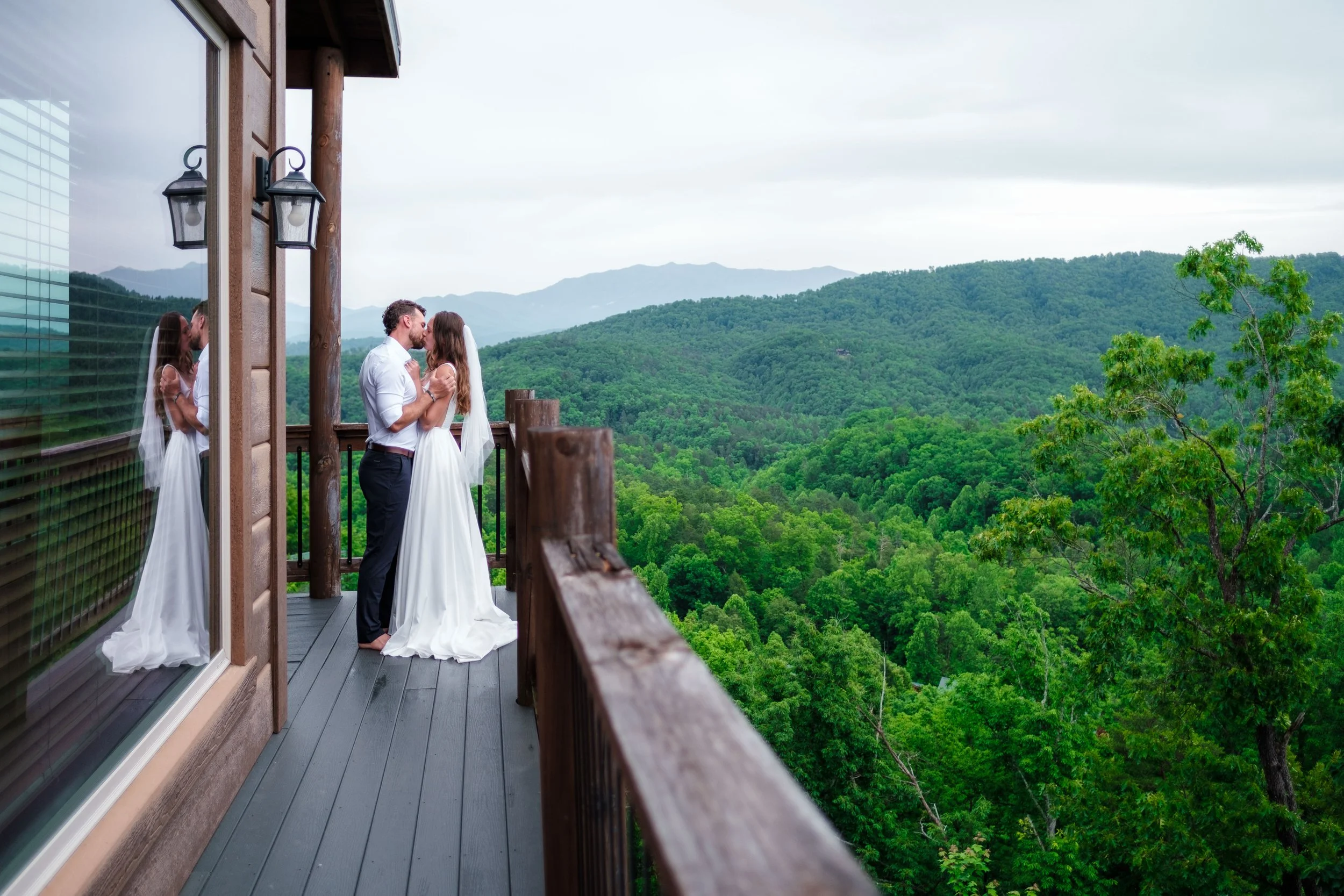A bride and groom sharing a kiss on a wooden balcony overlooking a lush green forest and rolling hills.