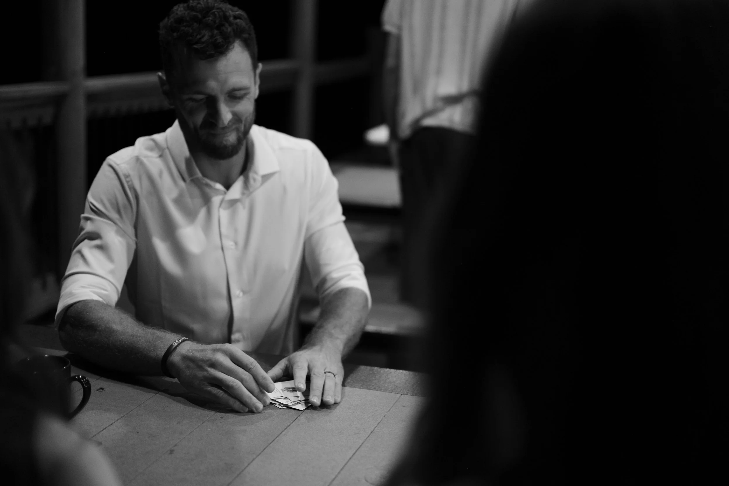 A man in a white shirt sitting at a table, playing cards, smiling, with a mug nearby, in a dimly lit room.