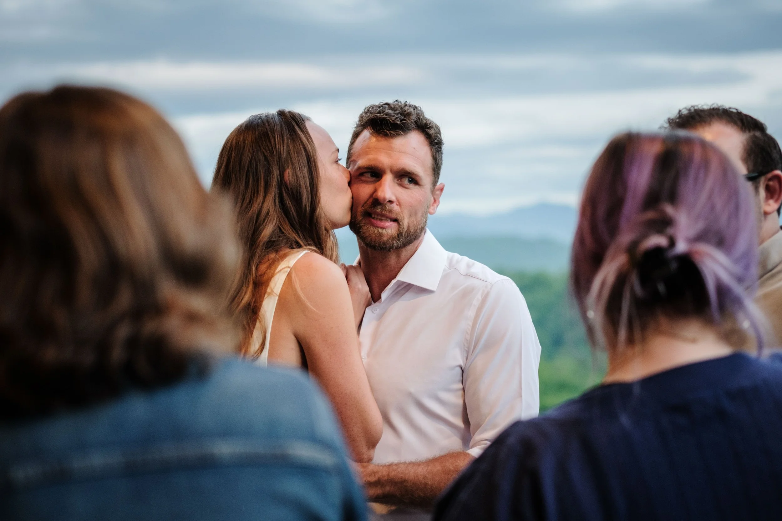 A woman kissing a man on the cheek during a ceremony outdoors, with the groom looking emotional, surrounded by guests with blurred backgrounds of mountains and cloudy sky.