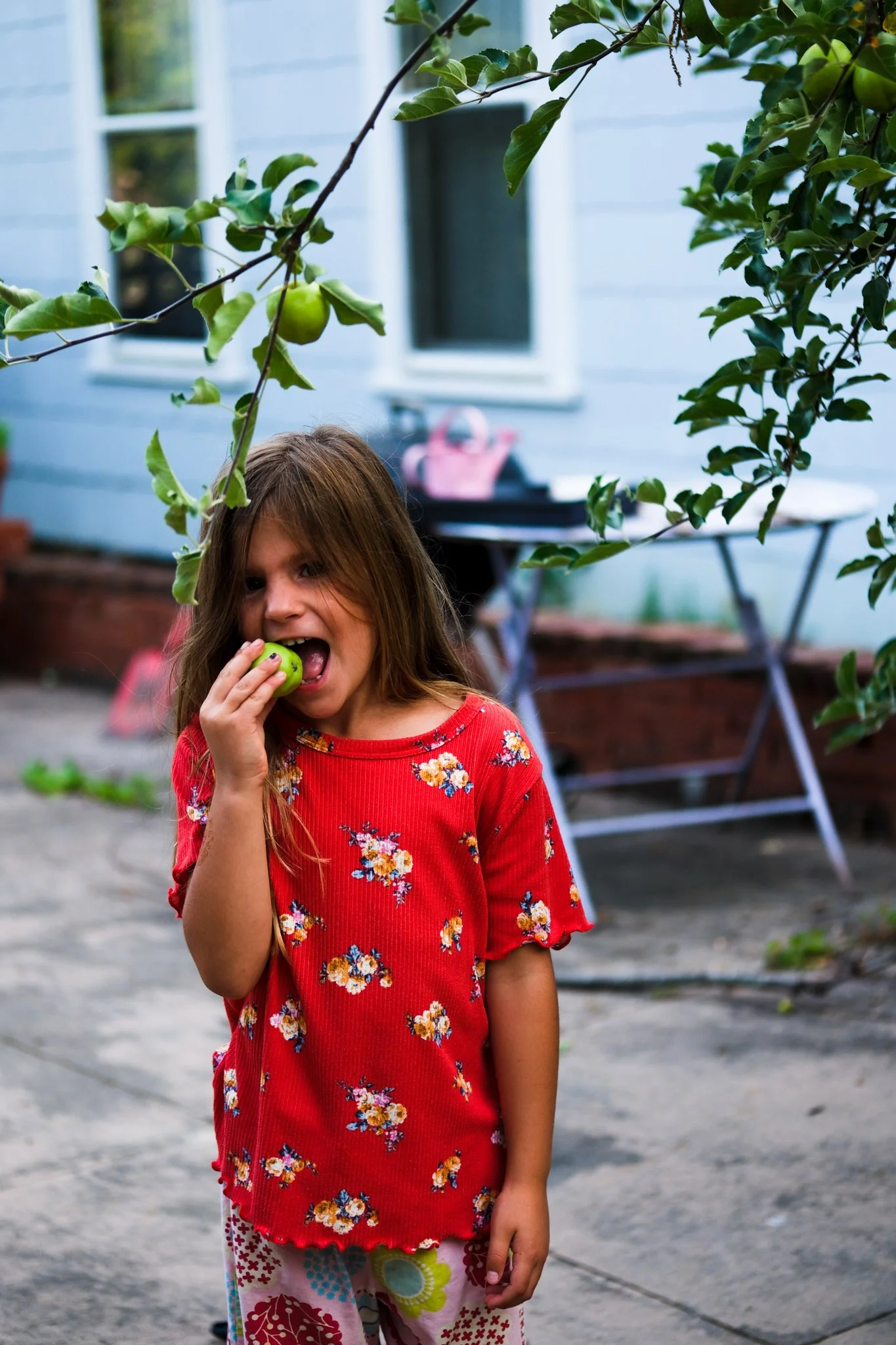 A young girl with long brown hair, wearing a red floral shirt and patterned pants, is outdoors, eating a green apple. She is standing in a backyard with a blue house, trees, and gardening equipment in the background.