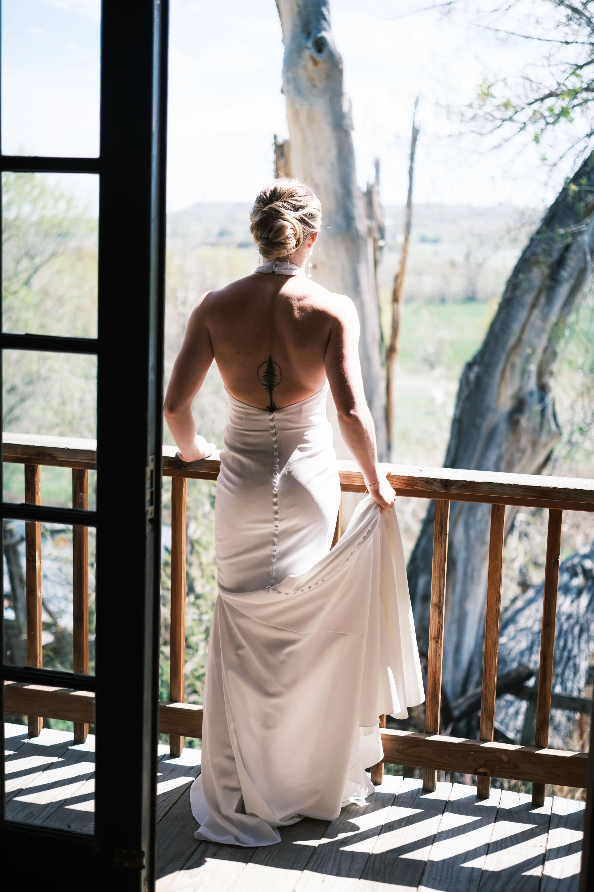 A woman in a backless white dress with buttons down the back stands on a wooden balcony, holding her dress with one hand, overlooking a tree outside.