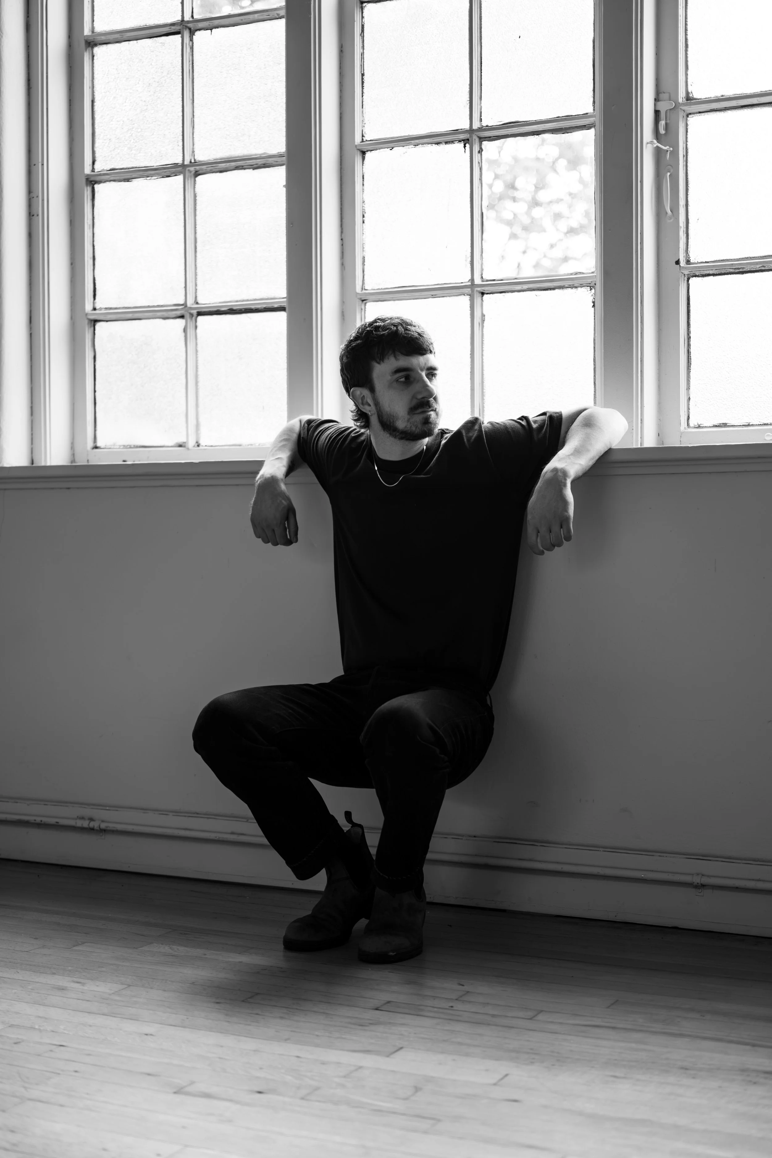 A man in black clothing sitting on the floor, leaning against a wall beneath a large window, looking thoughtfully outside.