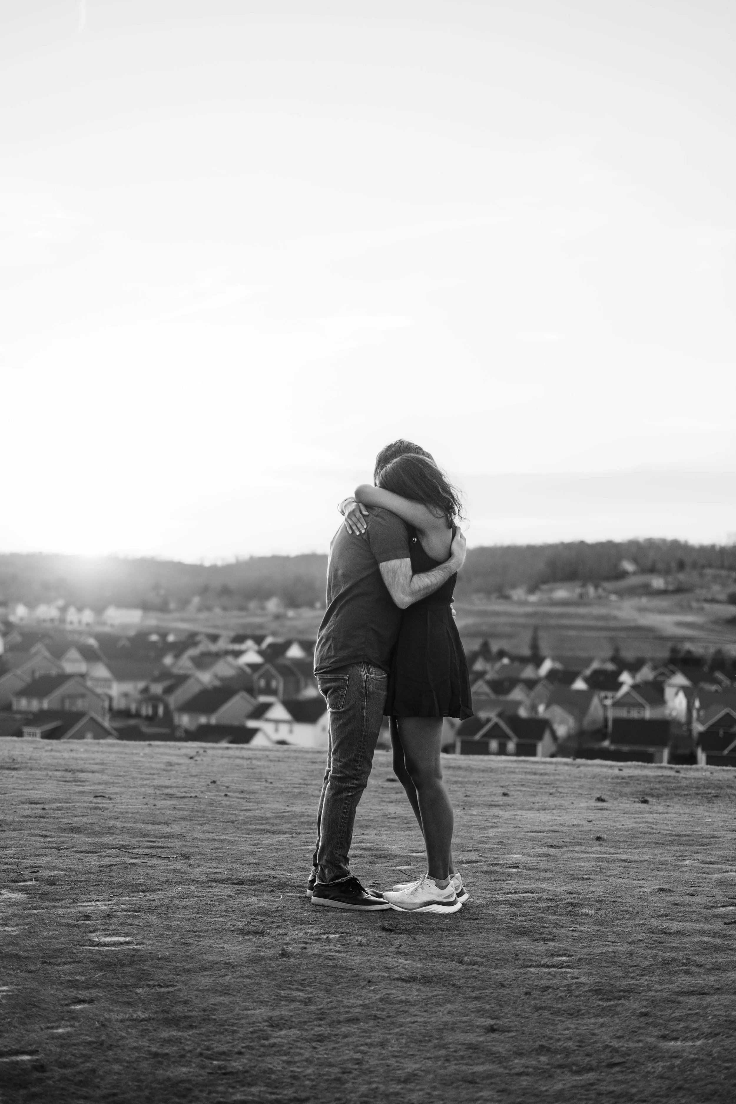 A black and white photo of a couple embracing outdoors on a hilltop with a view of a small town below at sunset.