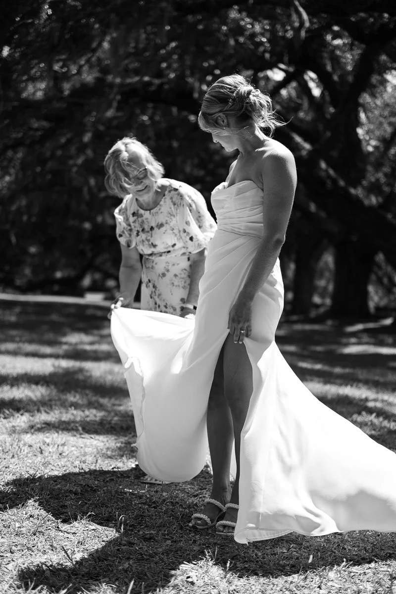 Black and white photo of a bride in a wedding gown outdoors, adjusting her dress, with an older woman assisting her, under a large tree.
