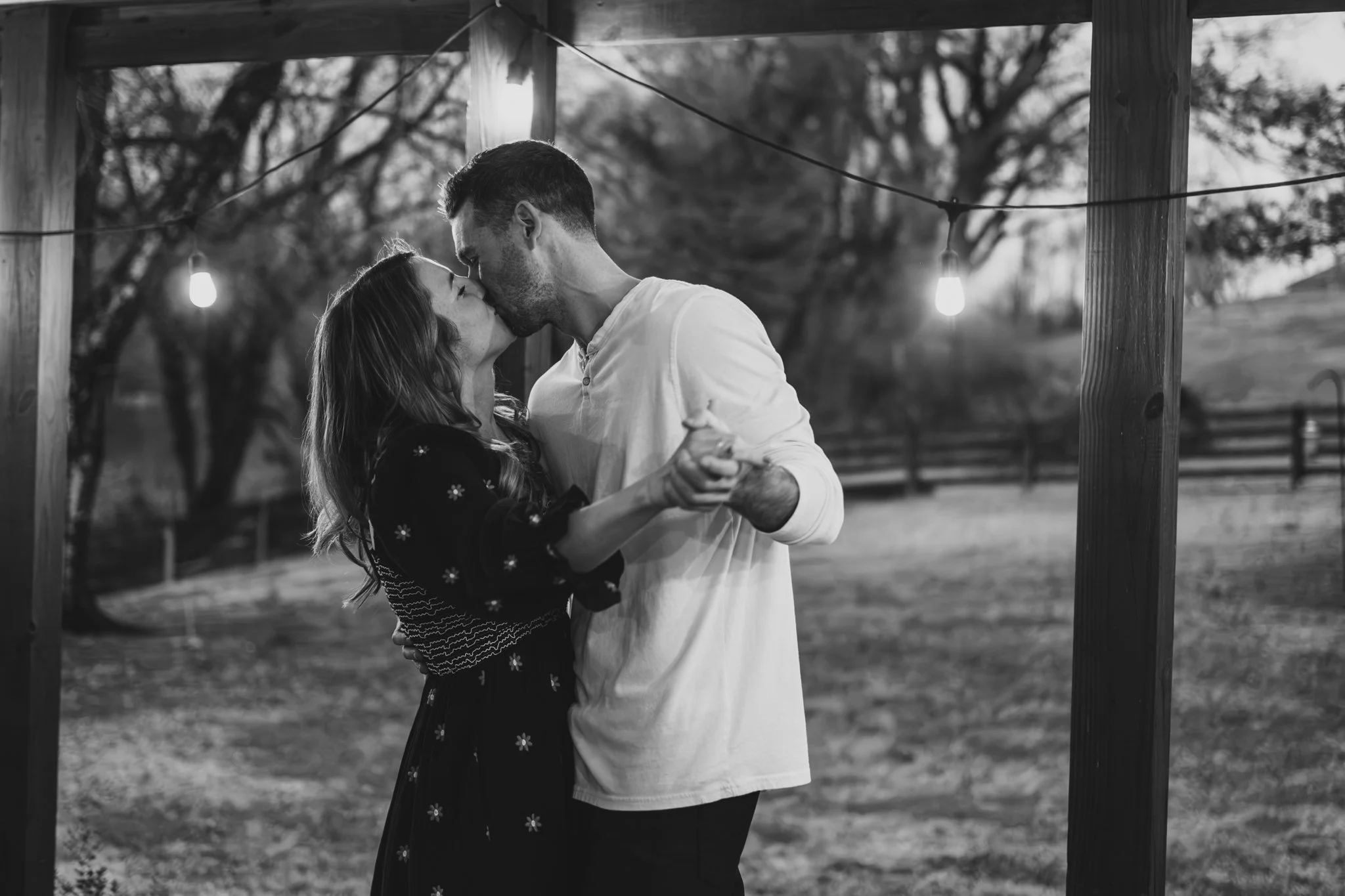 A black-and-white photo of a couple dancing and kissing outdoors at dusk under string lights, with trees and a wooden fence in the background.