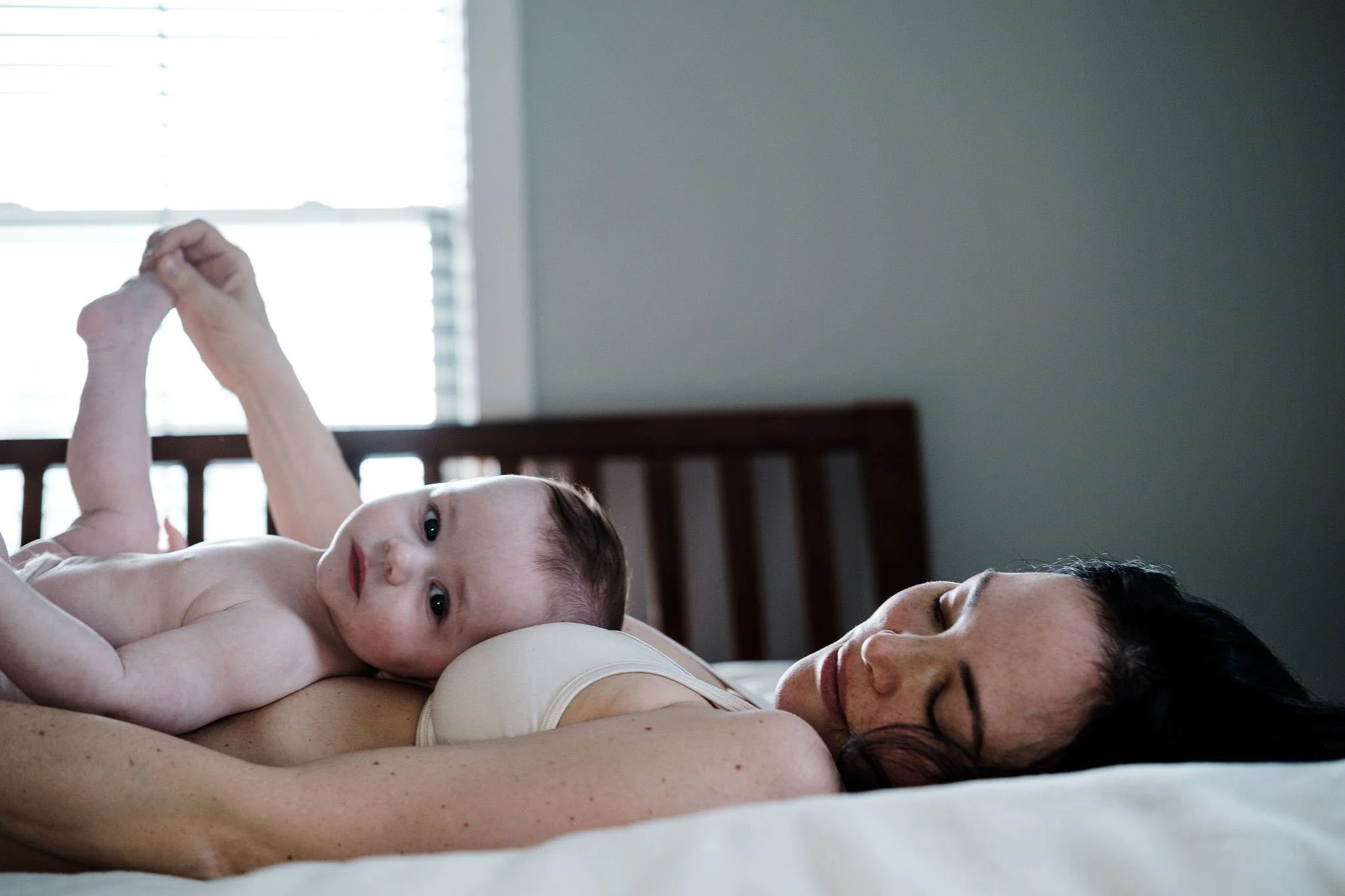 A woman lies on her side on a bed with a baby resting on her back, looking at the camera.