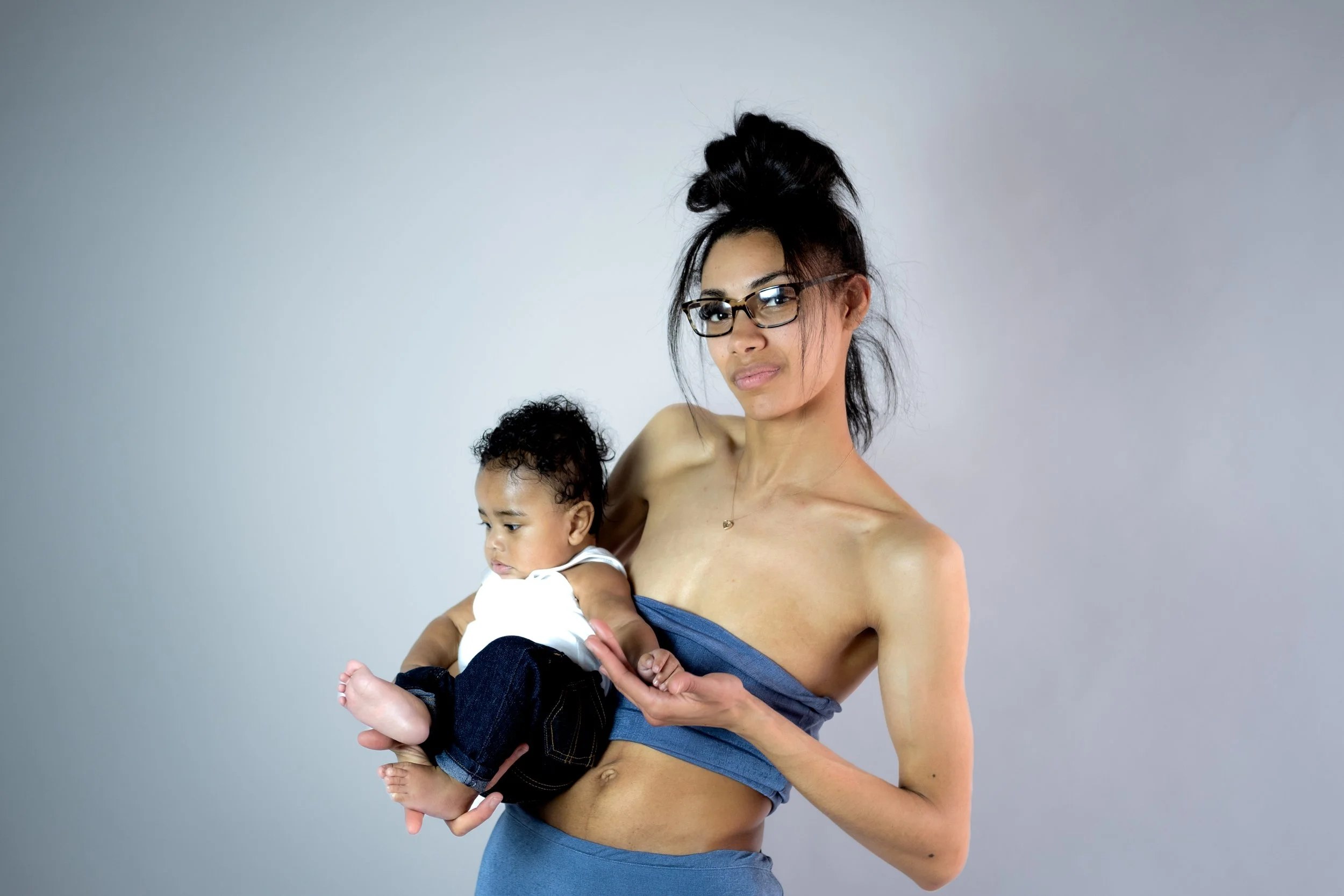 A woman with glasses holding a young child in her arms against a plain grey background.
