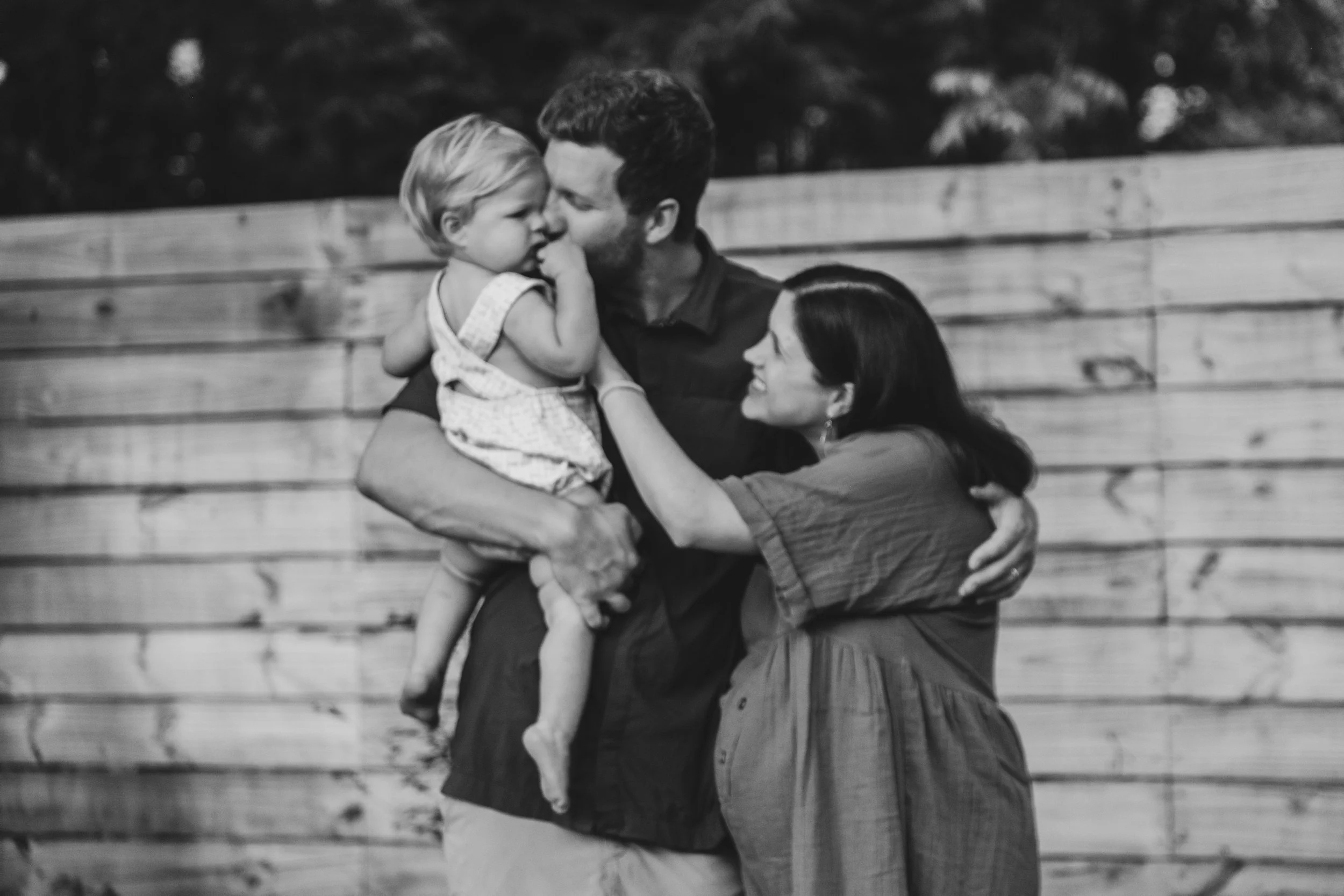A family of three sharing a tender moment outdoors. The father holds a young girl in his arms while the mother gently touches the girl’s face, all against a wooden fence backdrop.