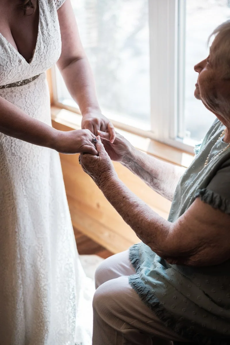 A woman in a wedding dress holding hands with an elderly woman sitting by a window.