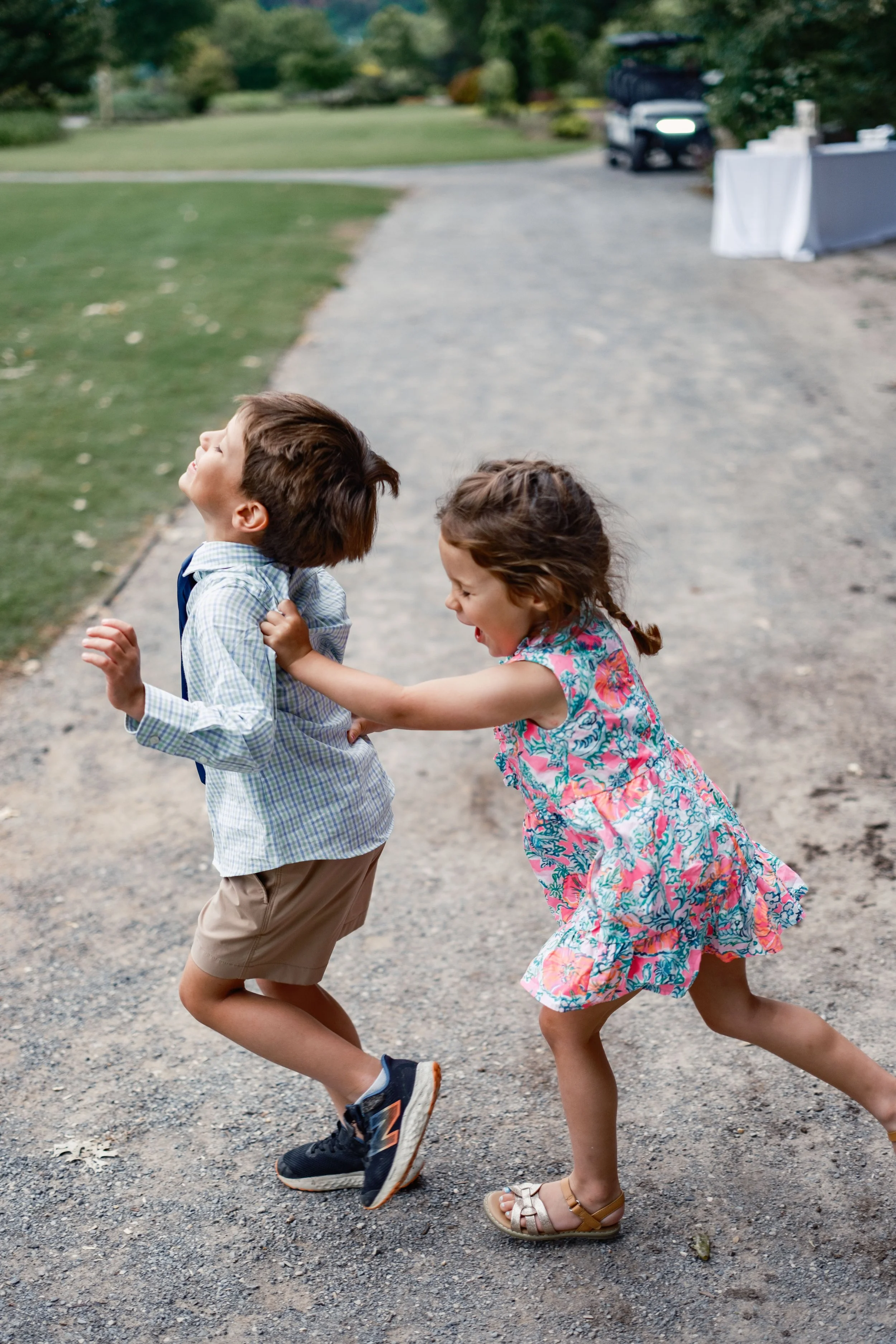 Two children, a boy and a girl, are playfully fighting and laughing on a gravel path outdoors, with greenery and parked cars in the background.