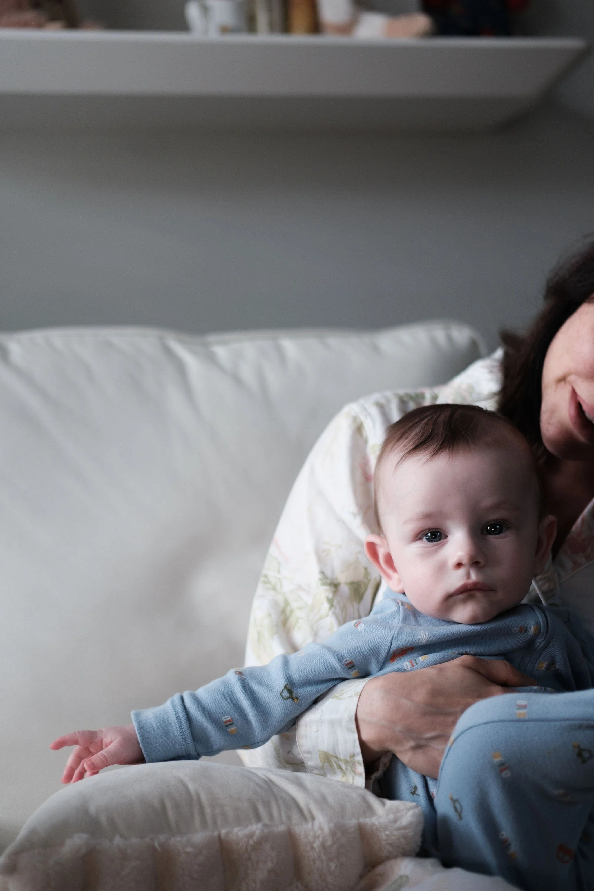 A baby with blue eyes and light brown hair sitting on a woman's lap, looking directly at the camera indoors. The woman, partially visible, is smiling and holding the baby gently.