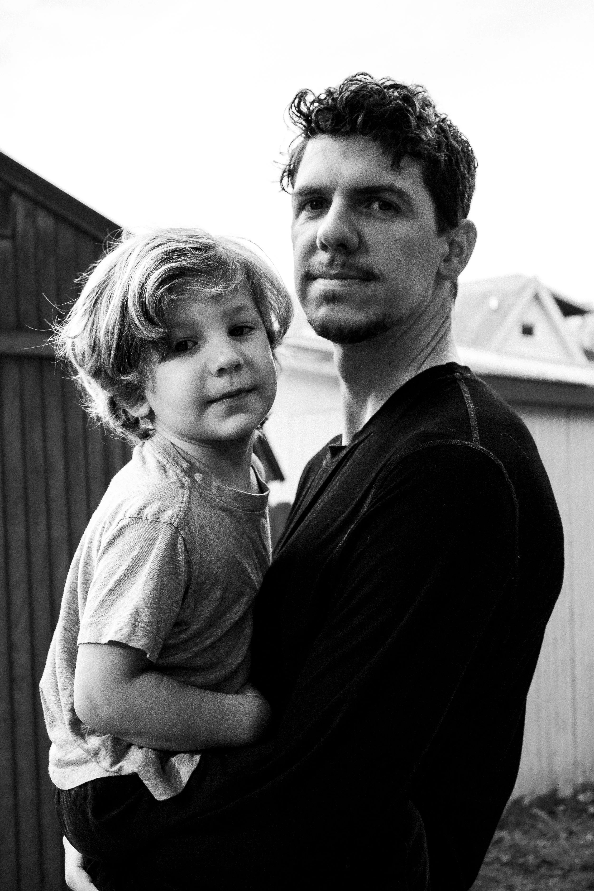 A man holding a young boy outdoors near a wooden fence, black and white photograph.