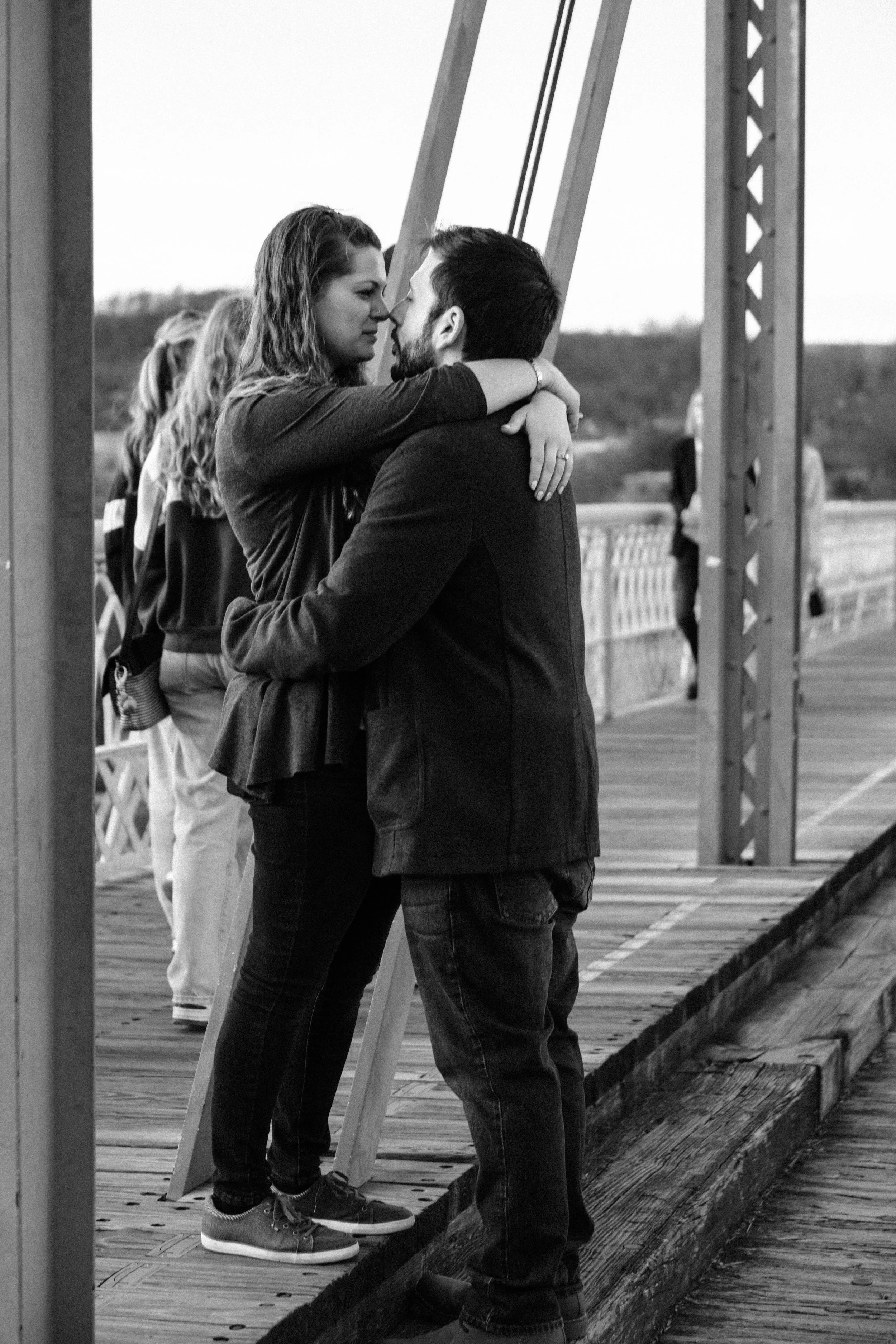 A black and white photo of a couple embracing on a wooden bridge, facing each other closely, with a woman sitting on the man's lap and their foreheads touching, outdoors with others in the background.