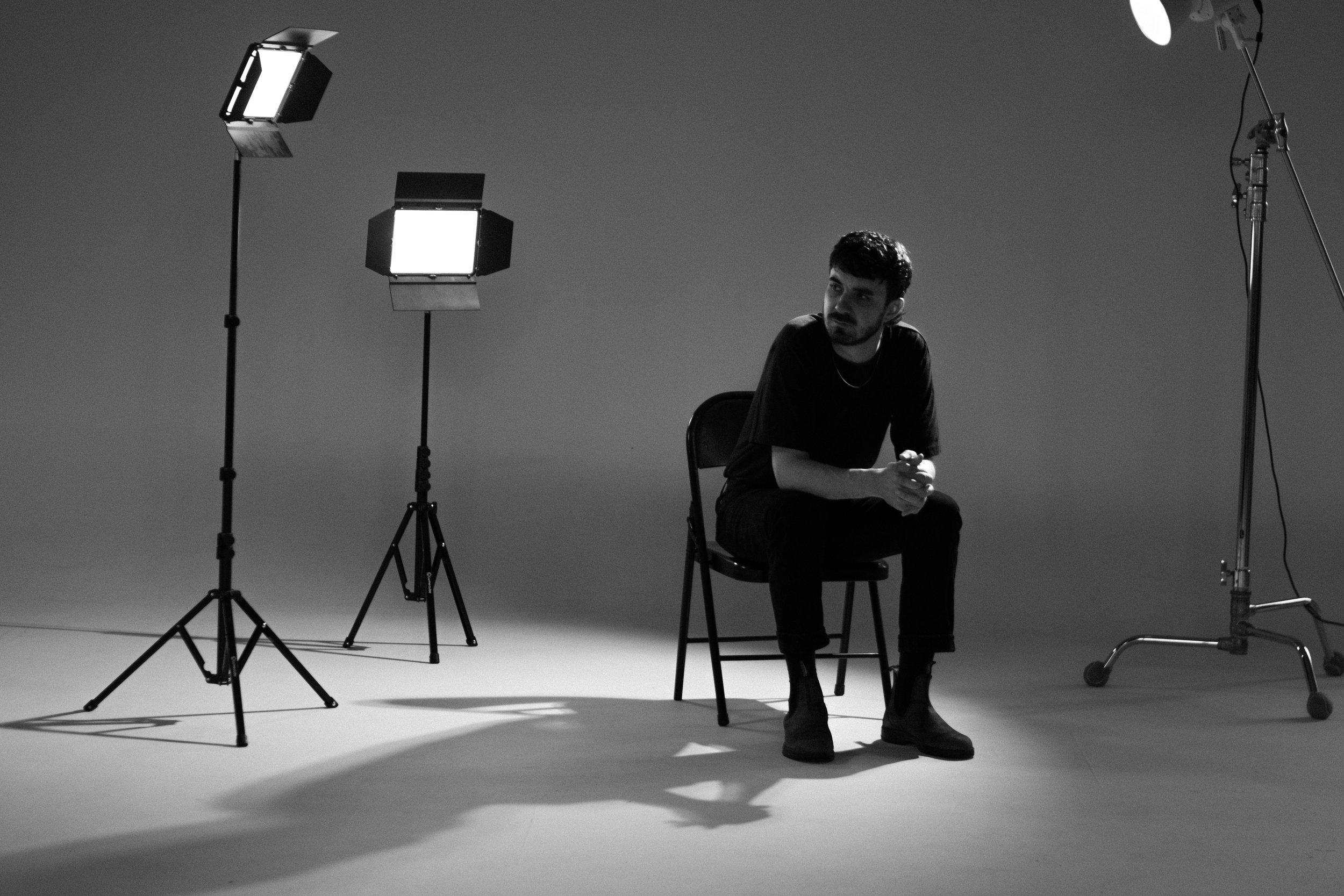 A man sitting on a chair in a photography studio with studio lights around him, in black and white.
