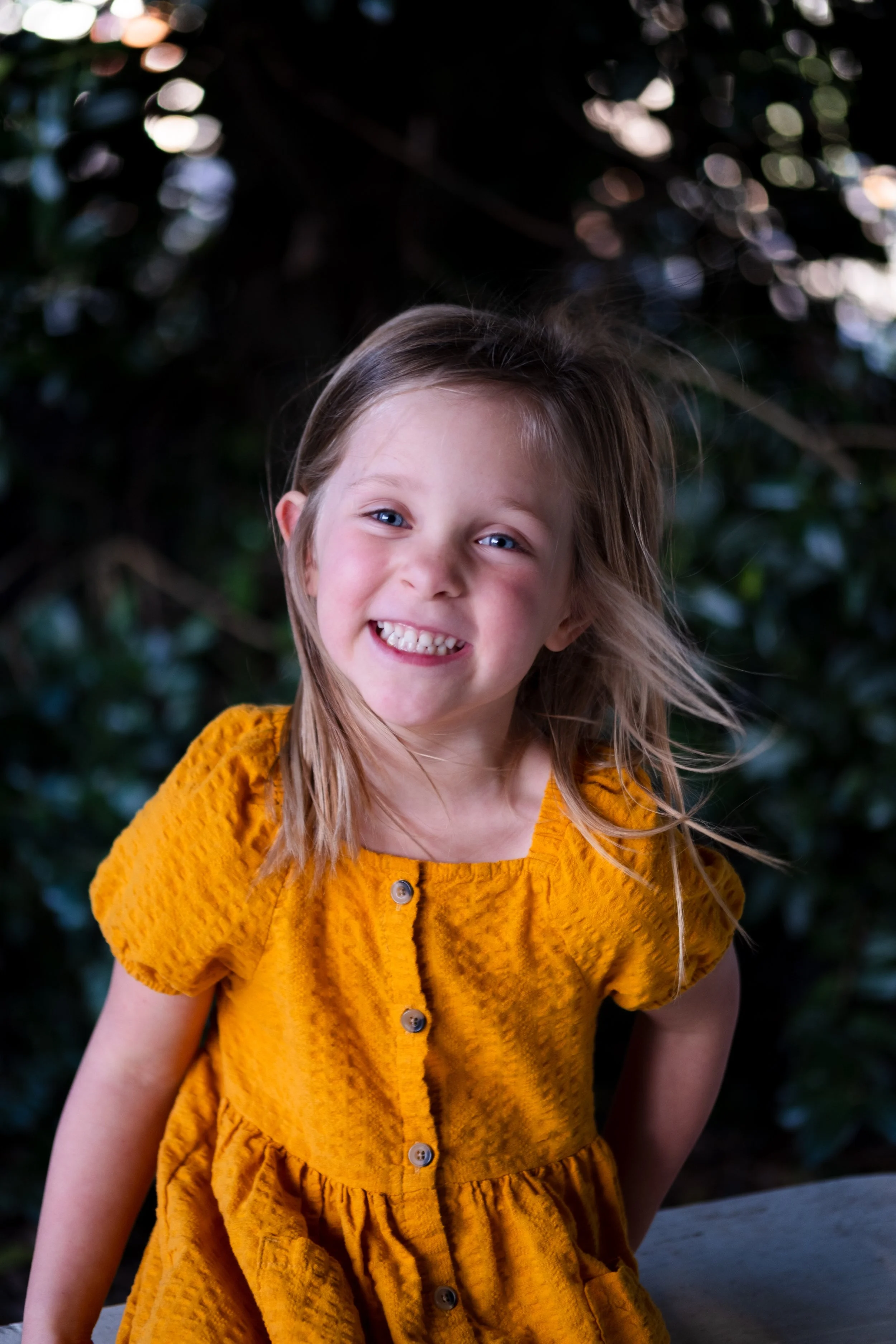 A young girl with blonde hair wearing a yellow dress, smiling and leaning forward outdoors.