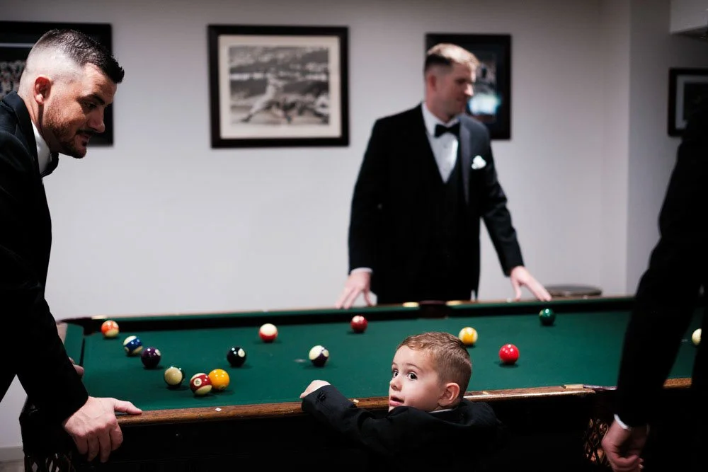 A young boy in formal attire looks surprised while leaning on a pool table, with three men in tuxedos around him in a room decorated with black and white framed photographs.
