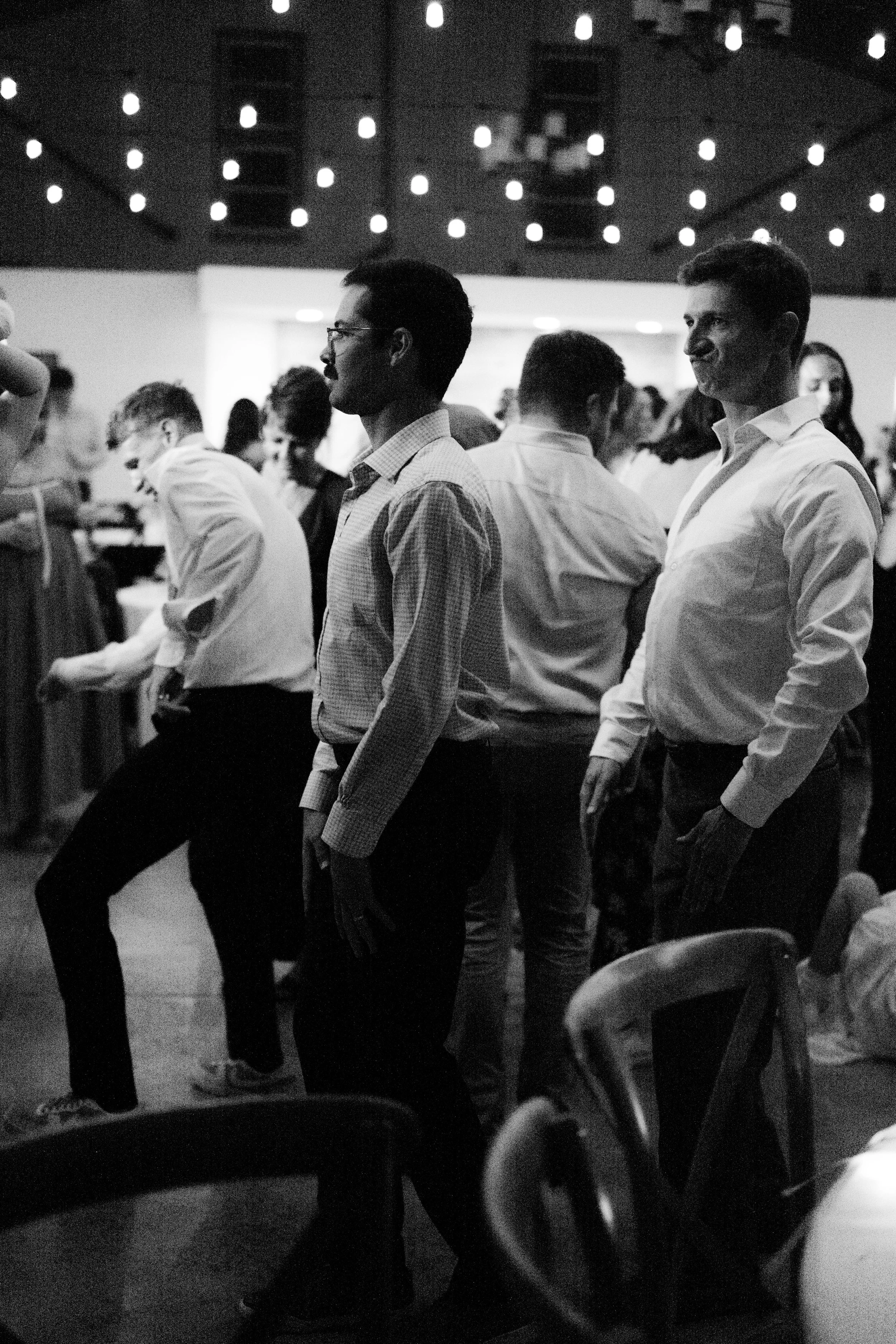 Black and white photo of people standing in a line or queue at an event, indoors with overhead string lights