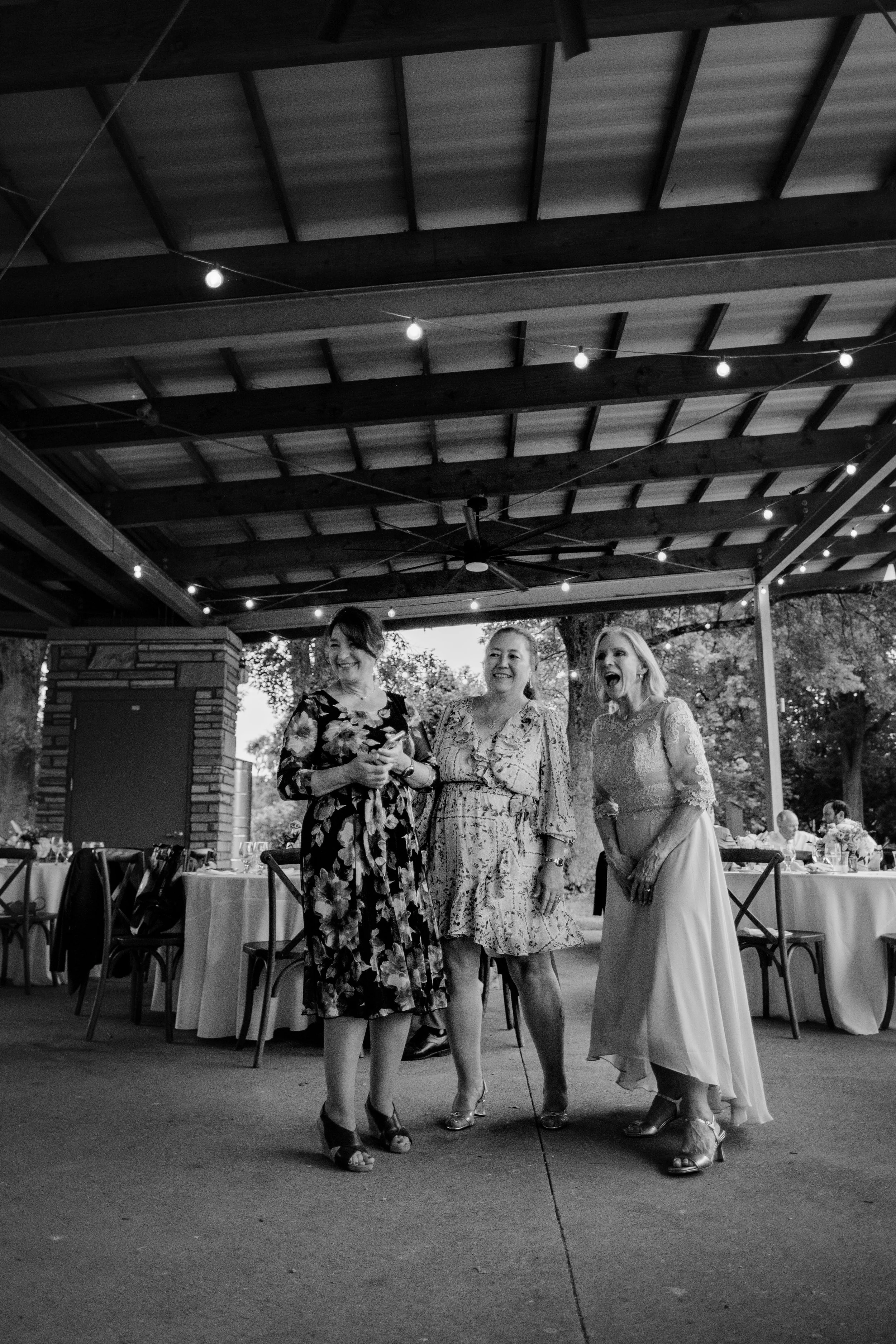 Three women at a social gathering, standing and smiling under a patio with string lights, dressed in formal attire.