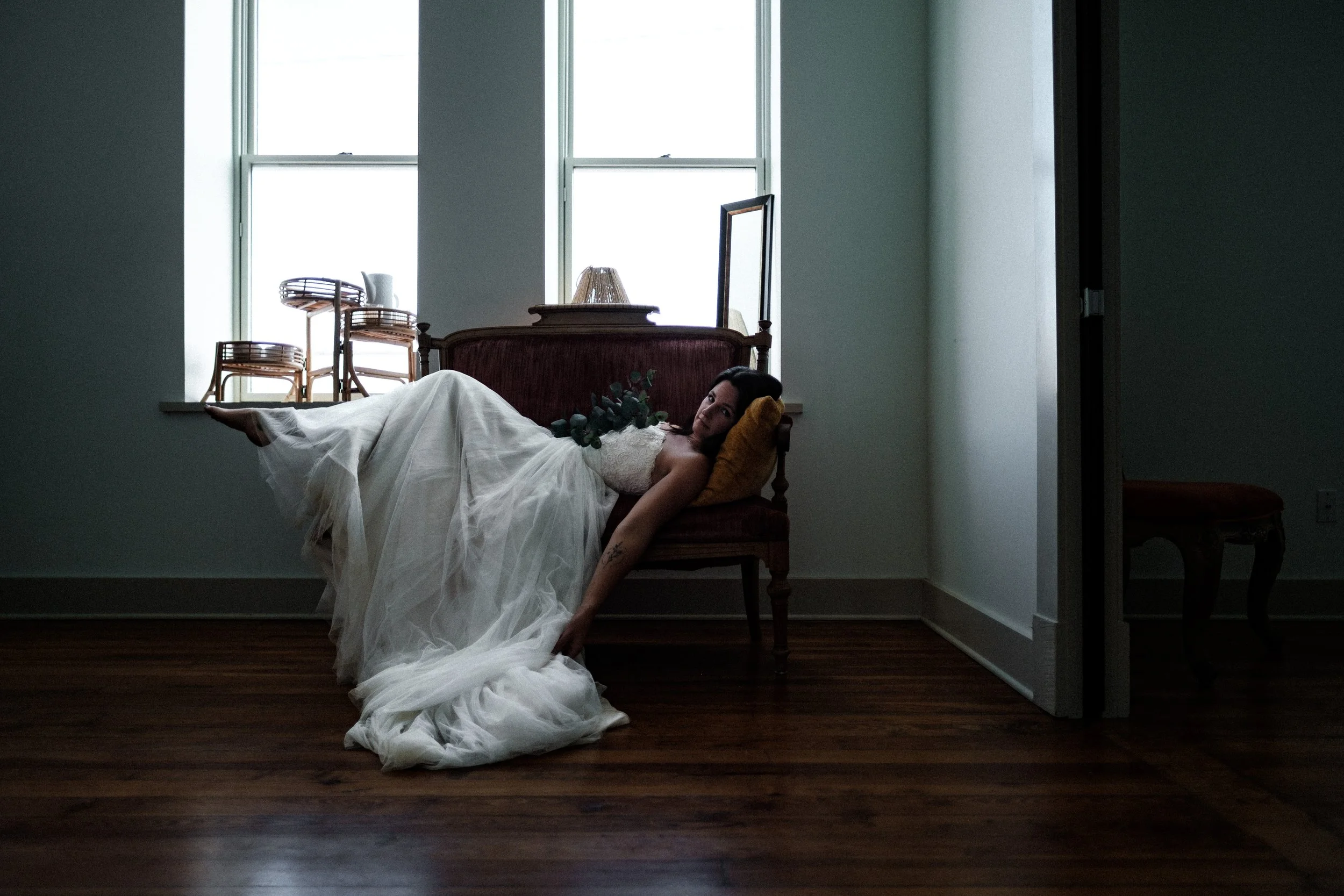 A woman in a wedding dress lying on a vintage sofa in a minimalist room with wooden floors and large window panes, holding a bridal bouquet and looking towards the camera.