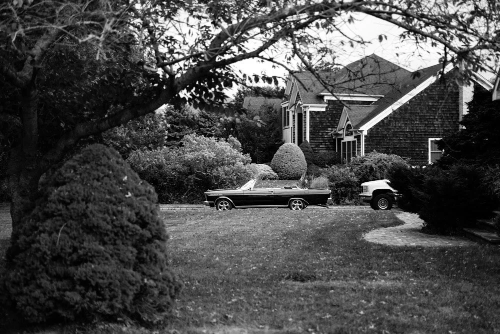A vintage car parked on a residential lawn with a large house and trees in the background.