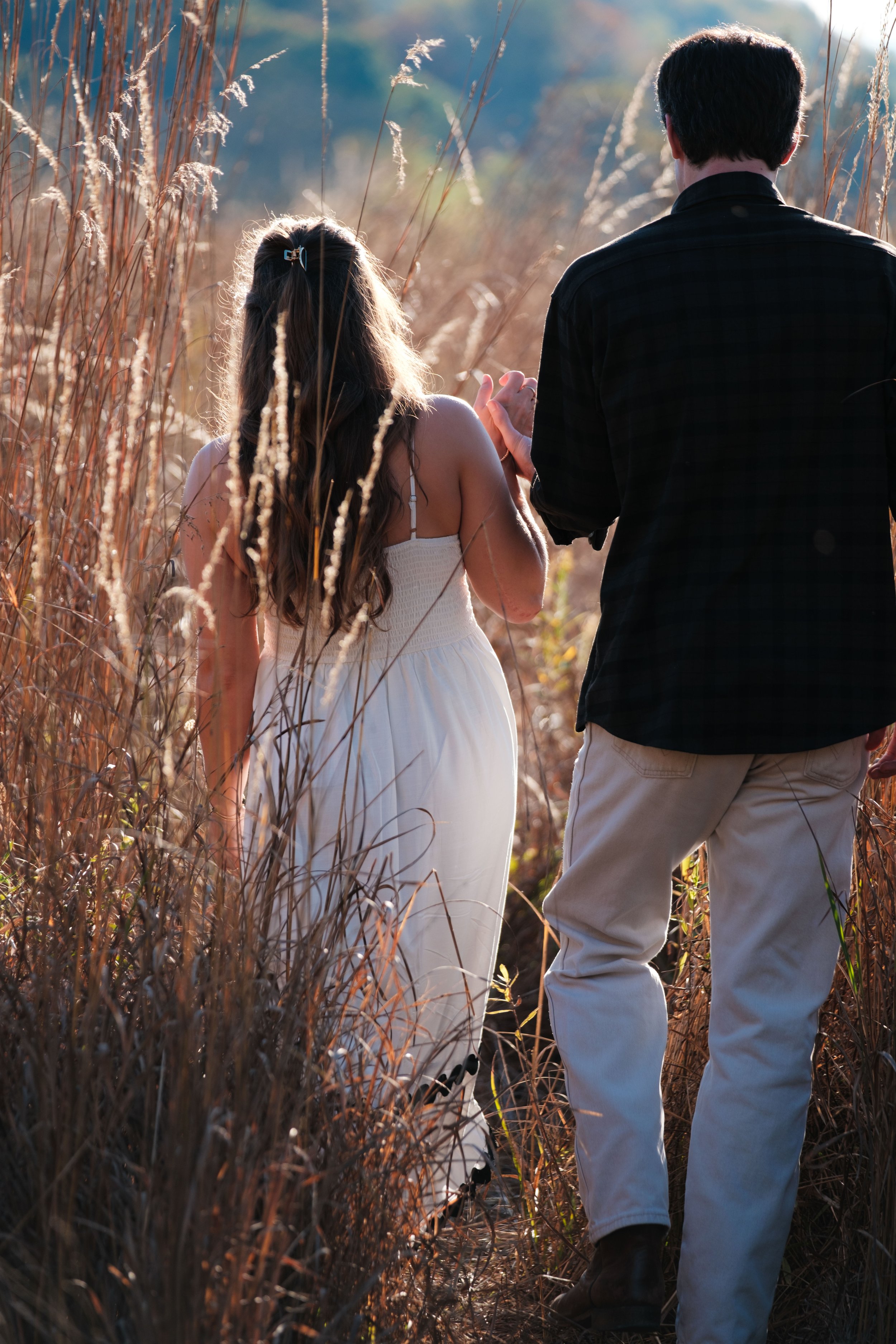 A couple walking through tall, dry grass in a field during sunset, holding hands.