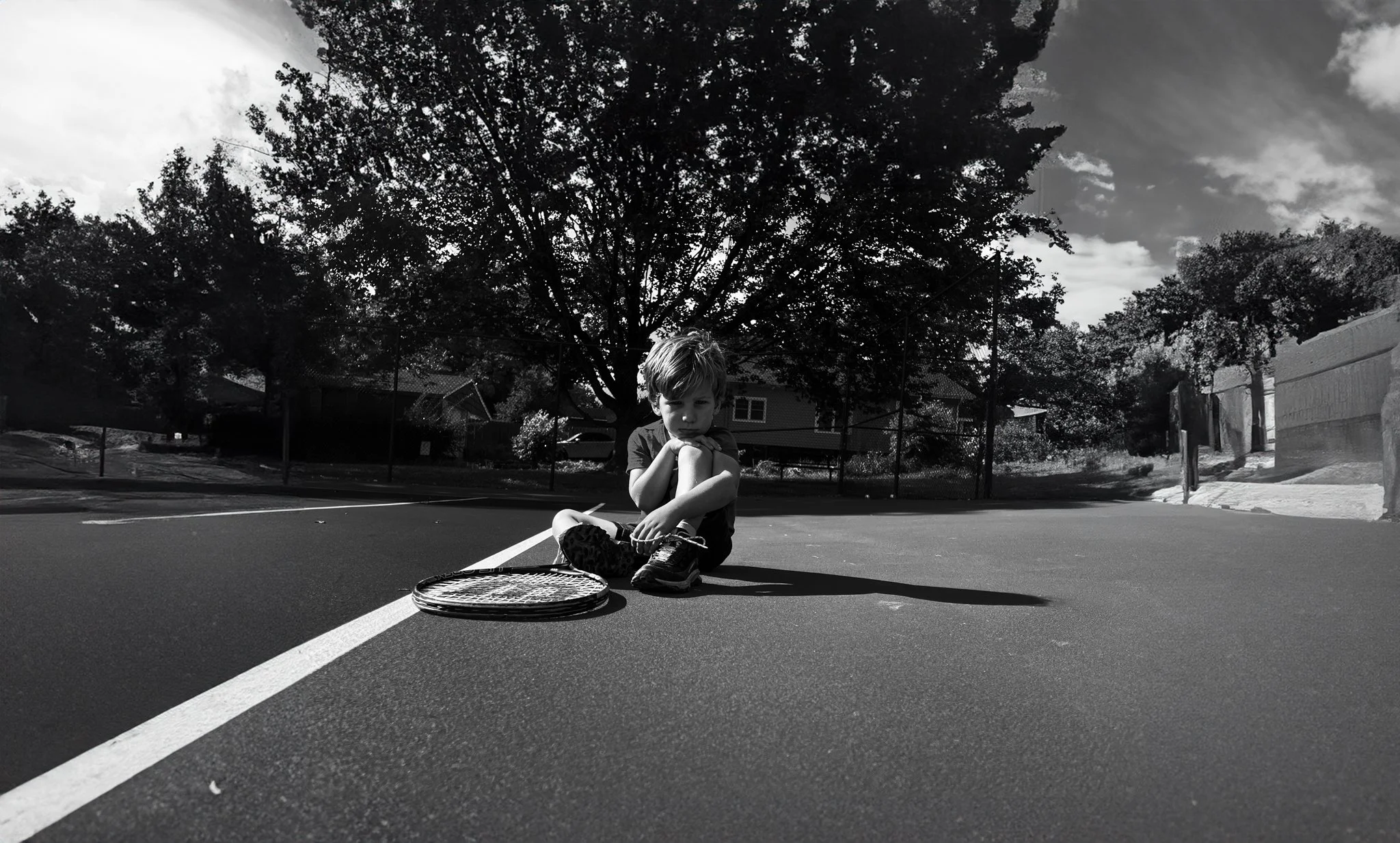 A young boy sitting on a basketball court holding his knees, with a tennis racket lying on the ground next to him. Trees and houses are visible in the background, and it is a partly cloudy day.