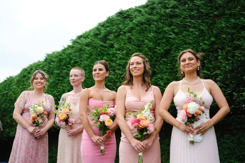 Five women in dresses holding bouquets, standing outdoors in front of a large green hedge, smiling for a photo.