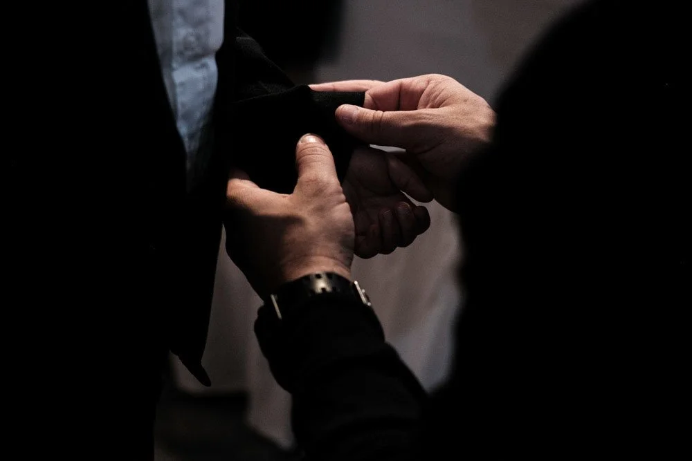 Two people shaking hands, one is wearing a black jacket with a watch, the other is wearing a dark suit and a white shirt.