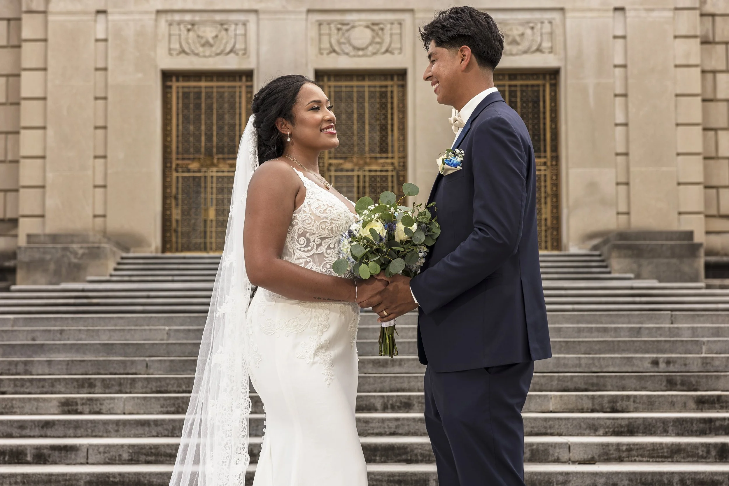Bride and groom standing on stairs outside a building, holding hands, looking at each other, smiling, wedding attire, wedding bouquet, bride in white dress with lace details, groom in navy suit with boutonniere, formal setting.