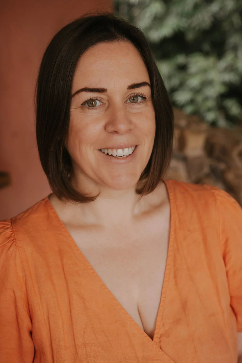 A woman with short dark brown hair and light skin, smiling, wearing an orange V-neck top, with a blurred background of greenery and a brick wall.