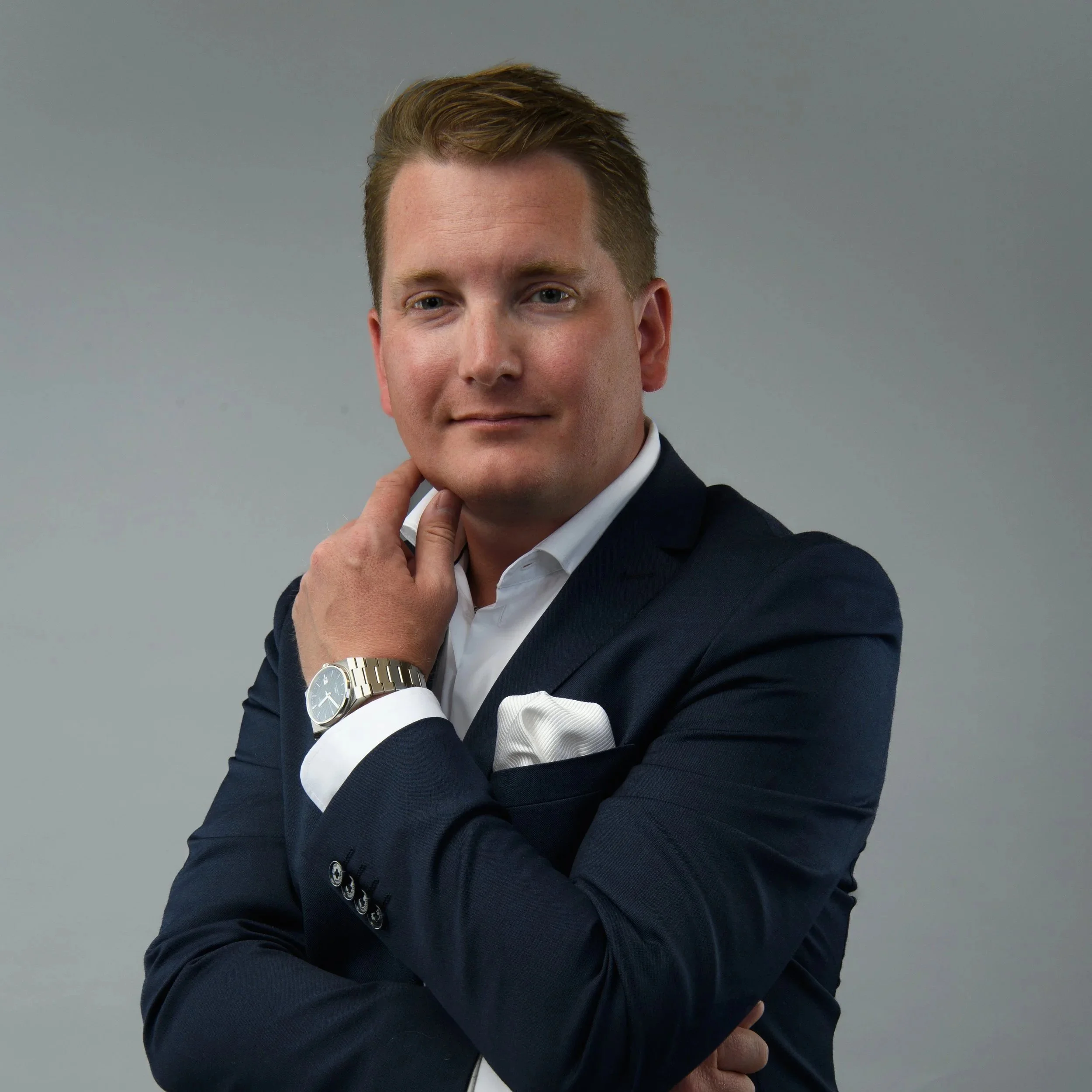 Ung man i formell kostym med mörkblå blazer och vit skjorta, poses, med armväcka, mot neutral grå bakgrund.