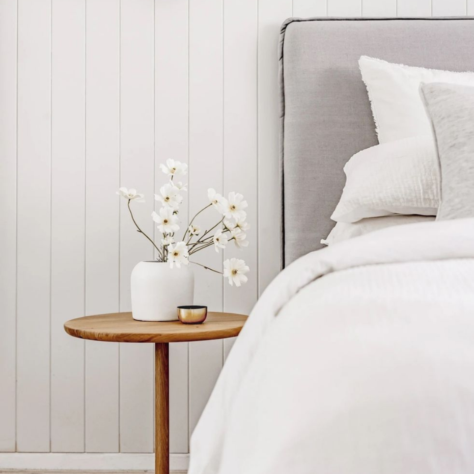 Close-up of a neatly made bed with white and light gray bedding, and a white pillow. Next to the bed, there is a small round wooden side table with a white vase containing white flowers and a small metallic bowl. The background features vertical white wooden paneling.