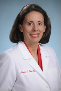 Portrait of a female doctor, Jeannene Dieter, wearing a white lab coat with red embroidery, smiling against a blue background.