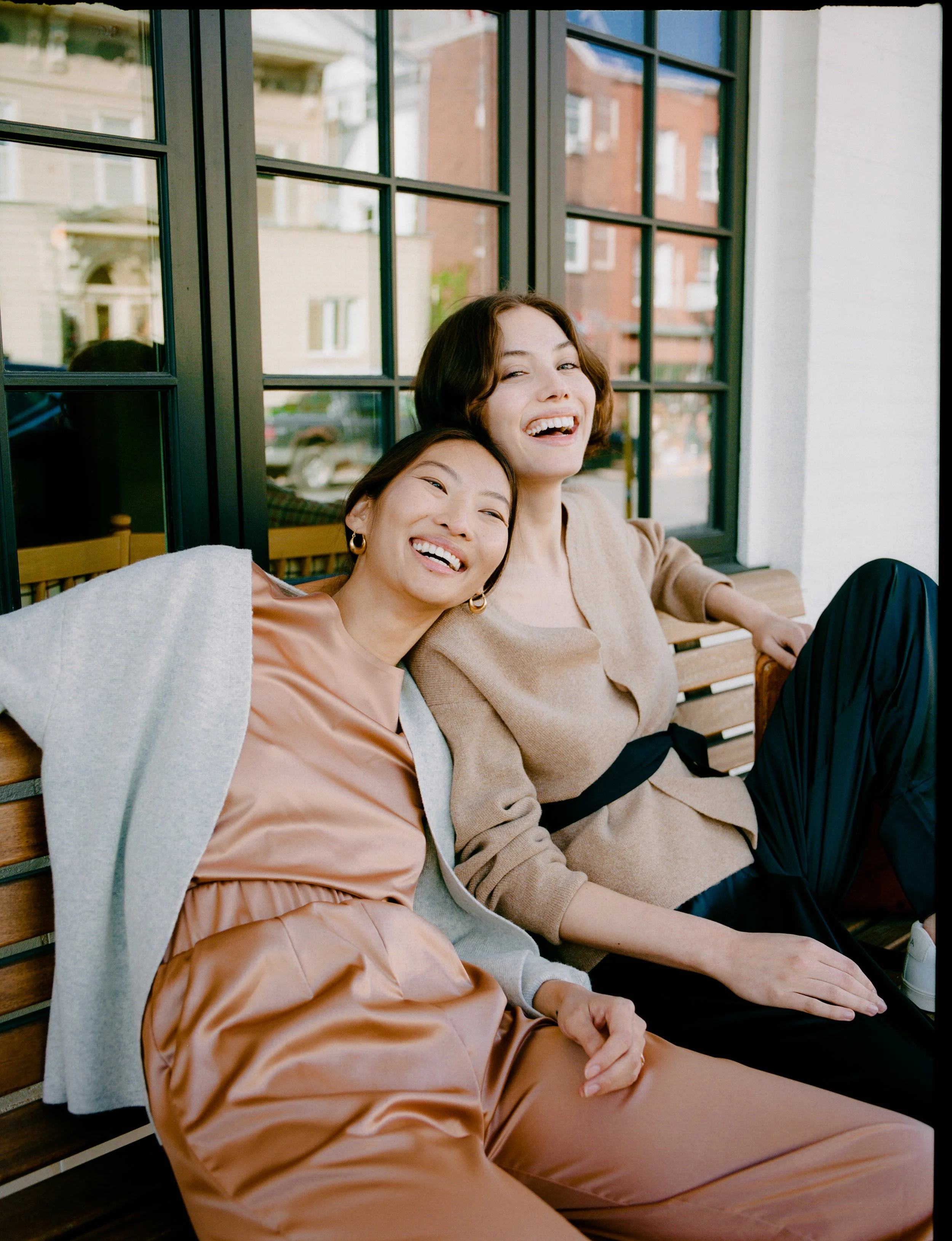 Two women sitting on a wooden bench, smiling and enjoying each other's company, in front of a window with a view of buildings outside.