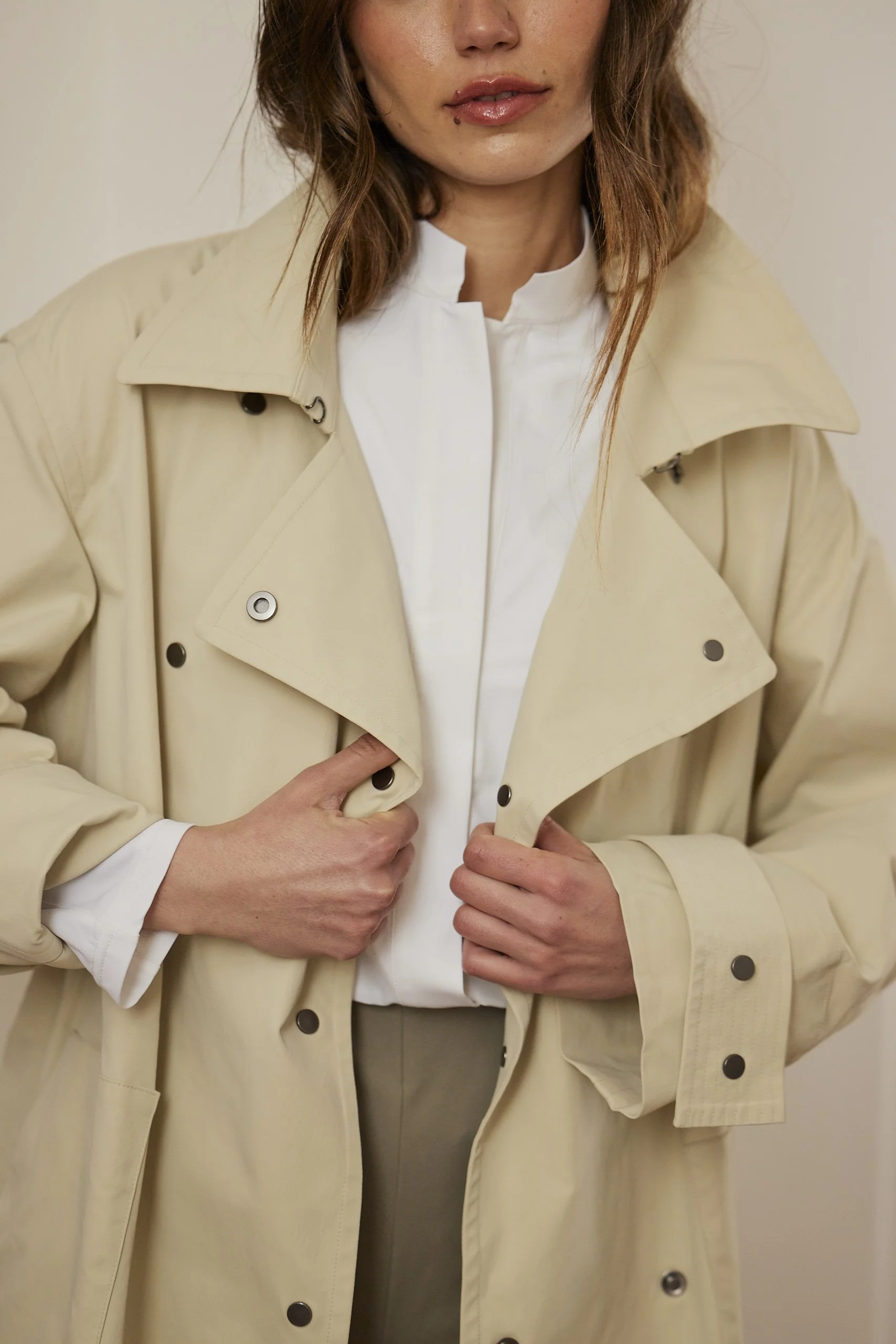 Close-up of a woman in a beige trench coat and white shirt, holding the coat closed with both hands.